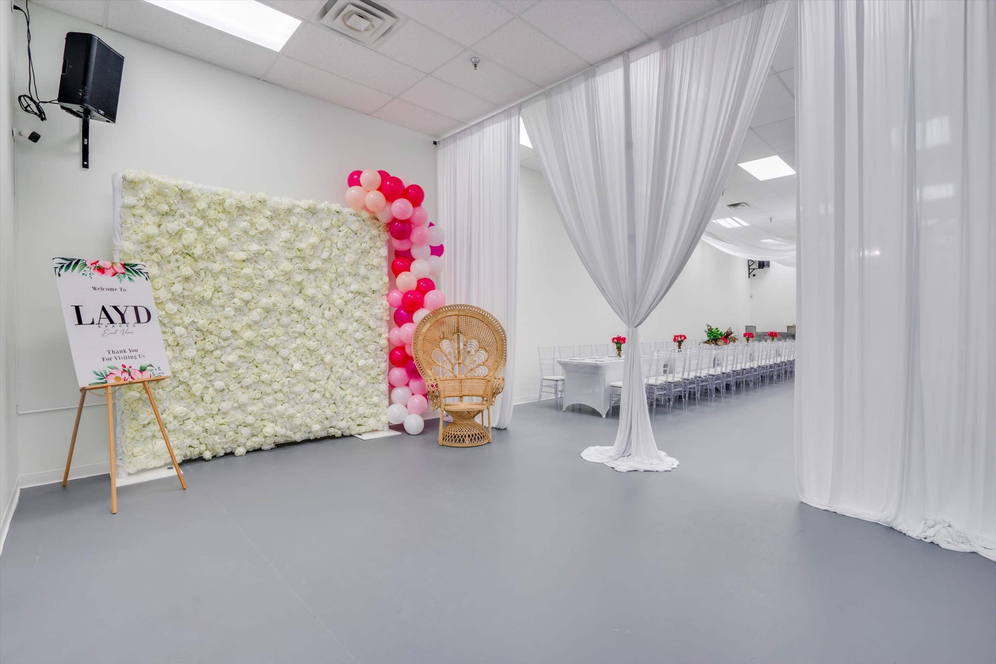 The image shows a spacious event venue featuring a floral wall decoration, a balloon arch, a decorative chair, and a long table set for a gathering.