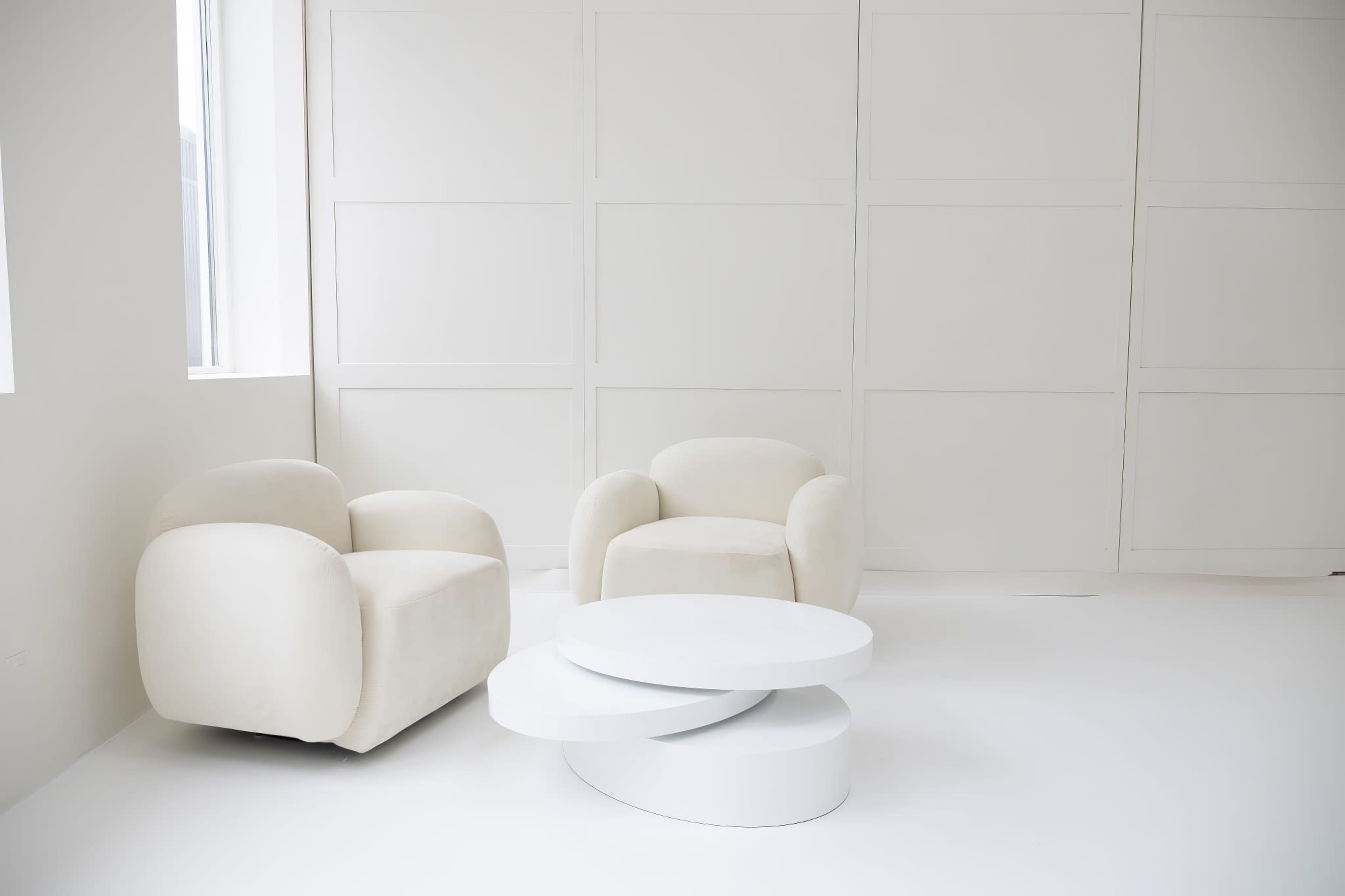 A minimalist interior with two cream-colored armchairs and a white modern coffee table against a plain wall.