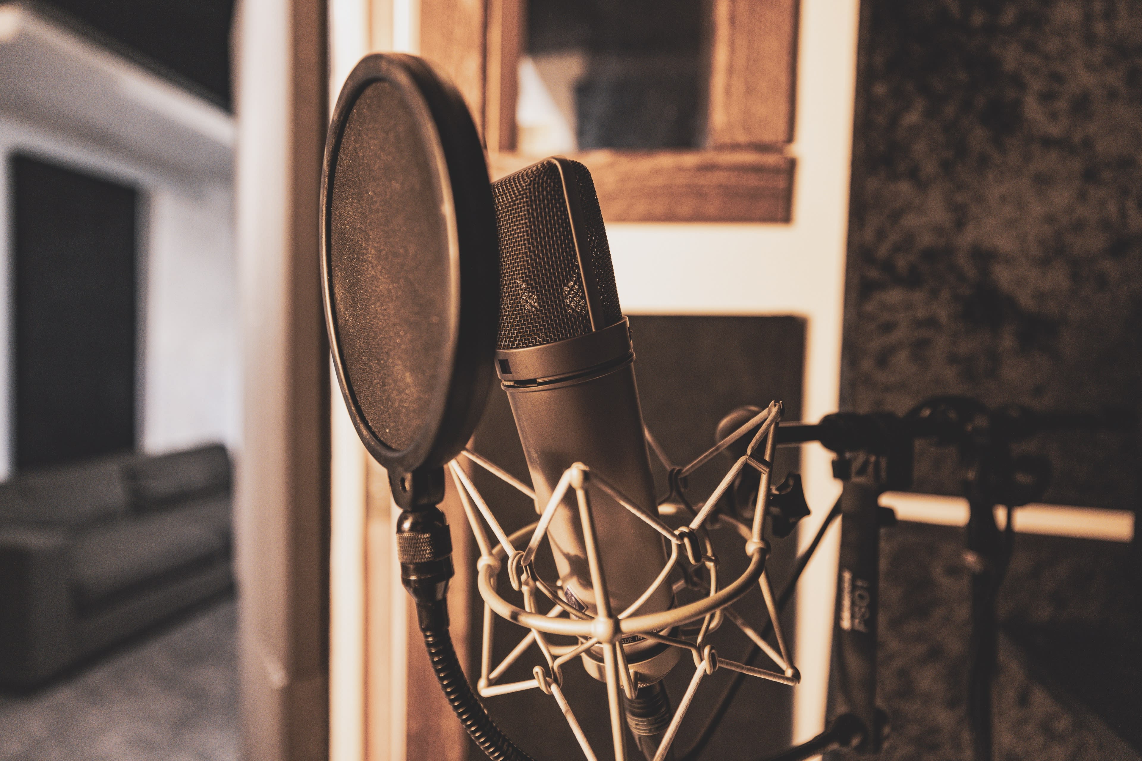 A microphone with a pop filter is positioned in a recording studio setting, with a blurred background showing a couch and studio elements.