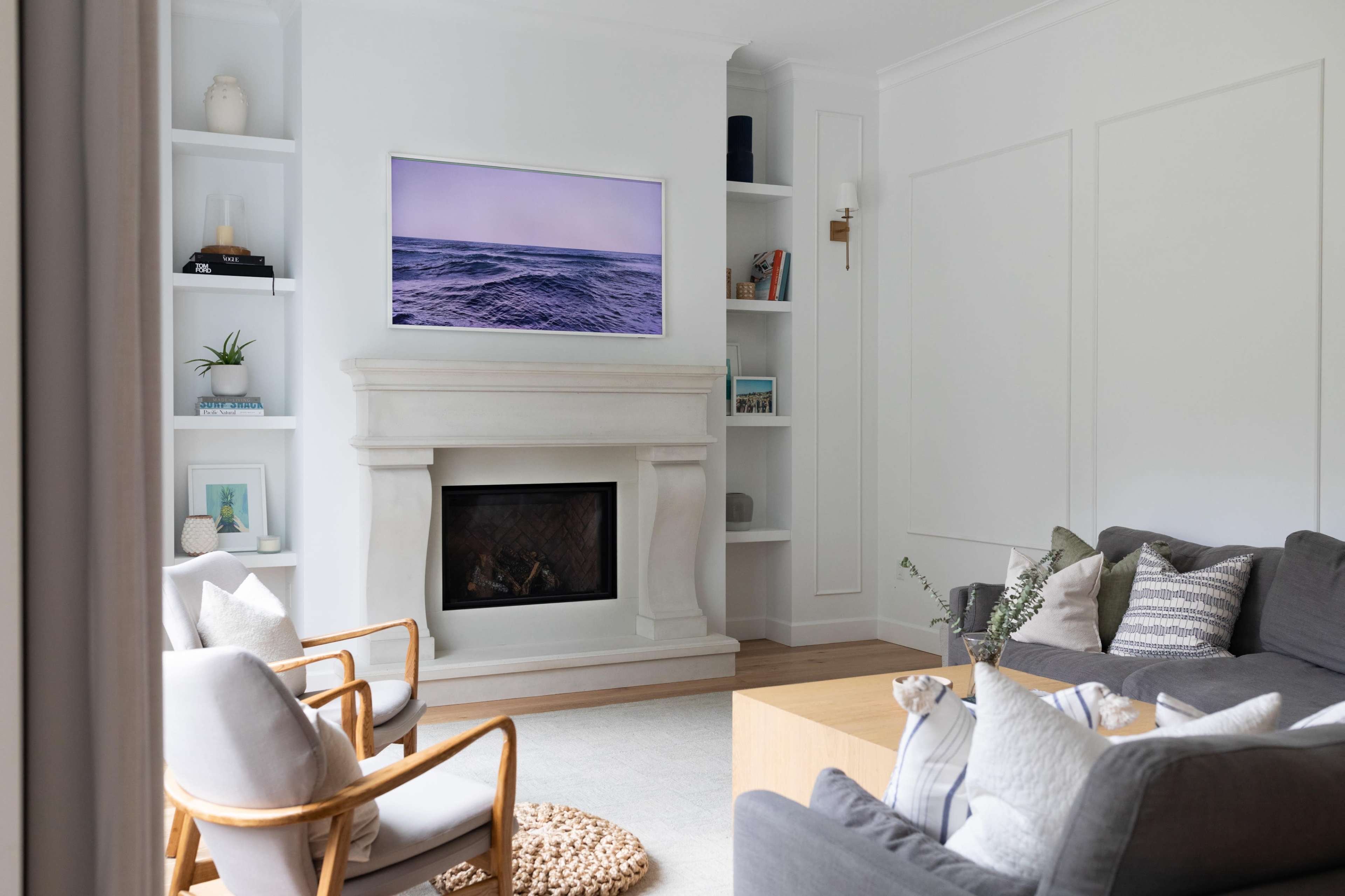A modern living room features a gray sofa and two wooden chairs facing a fireplace, with a large television mounted above the mantel, surrounded by built-in shelves displaying decorative items.