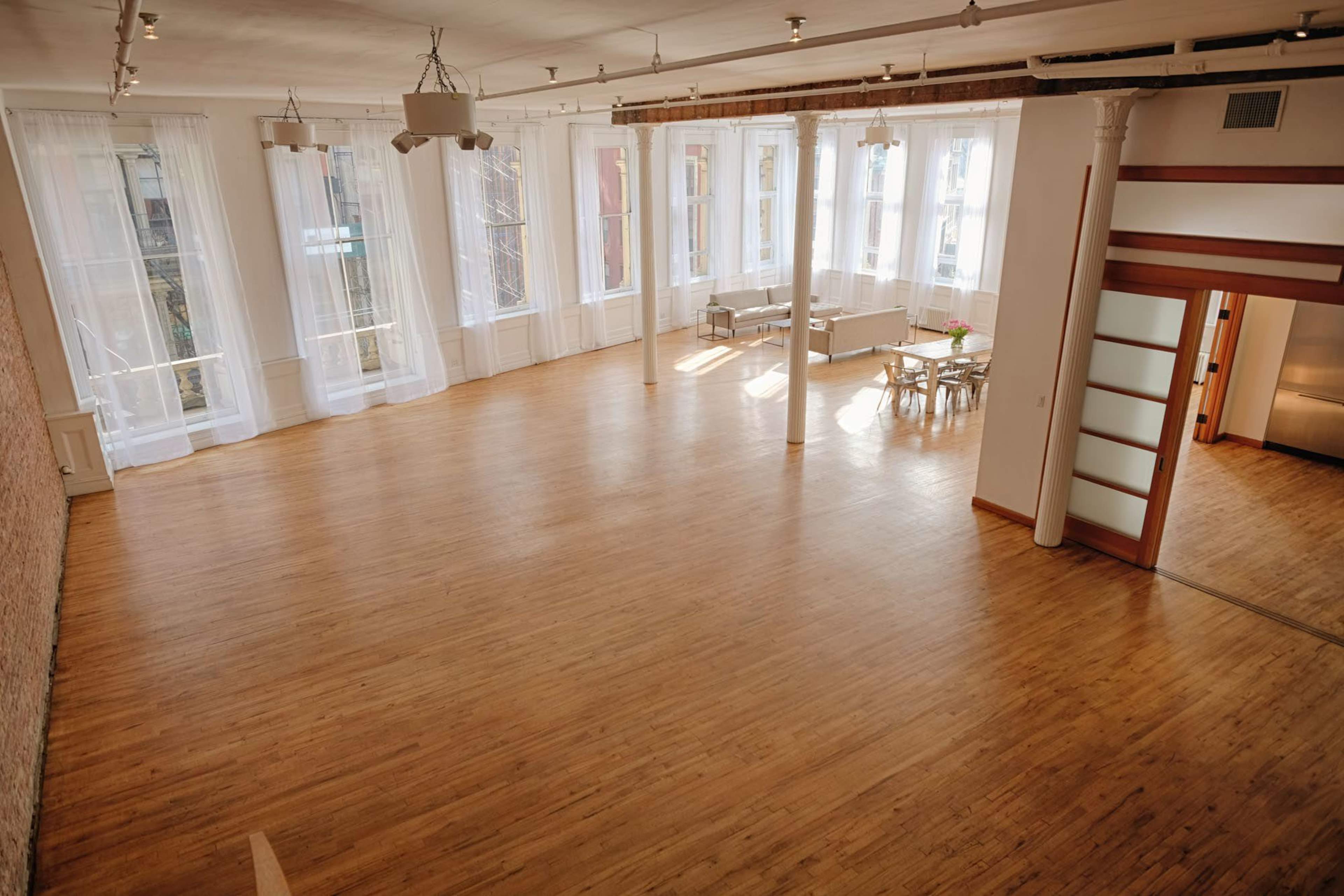 A spacious, well-lit room with large windows, wooden flooring, and a dining area in the corner.