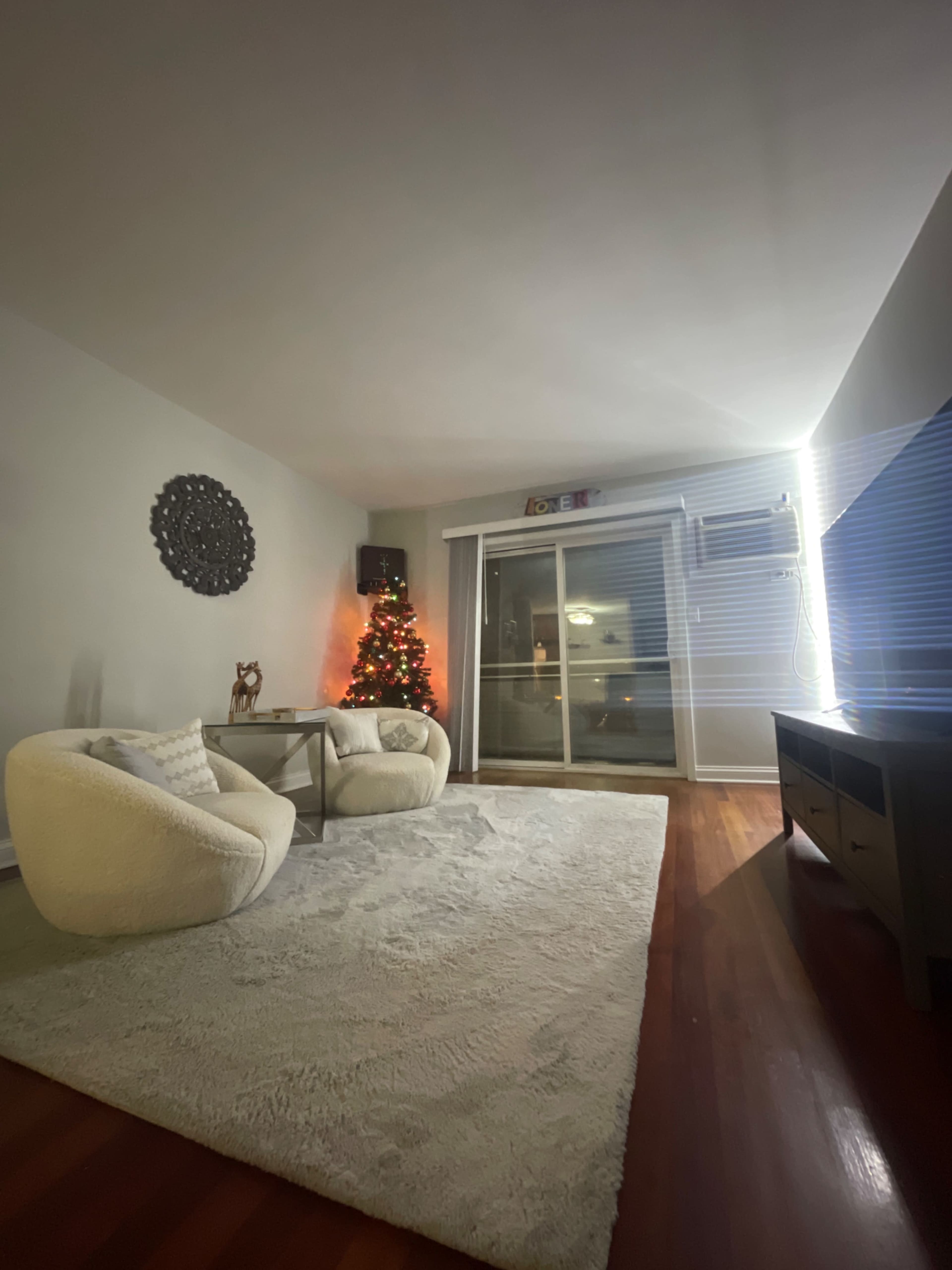 A cozy living room features a Christmas tree with lights, a round rug, and two armchairs facing a television and a glass door leading to a balcony.