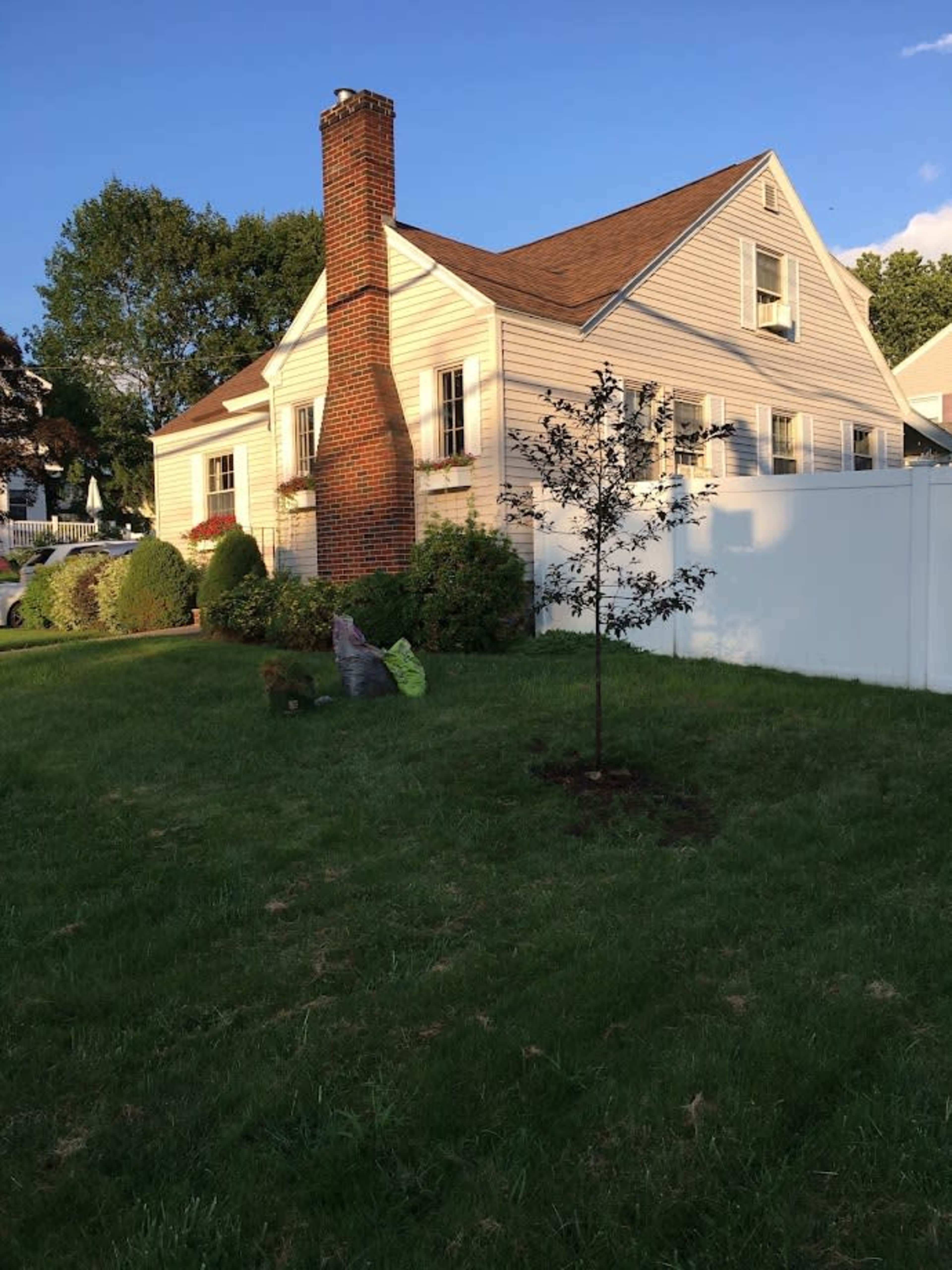 A single-story house with a brick chimney is positioned next to a fenced yard featuring a small tree and landscaped areas.