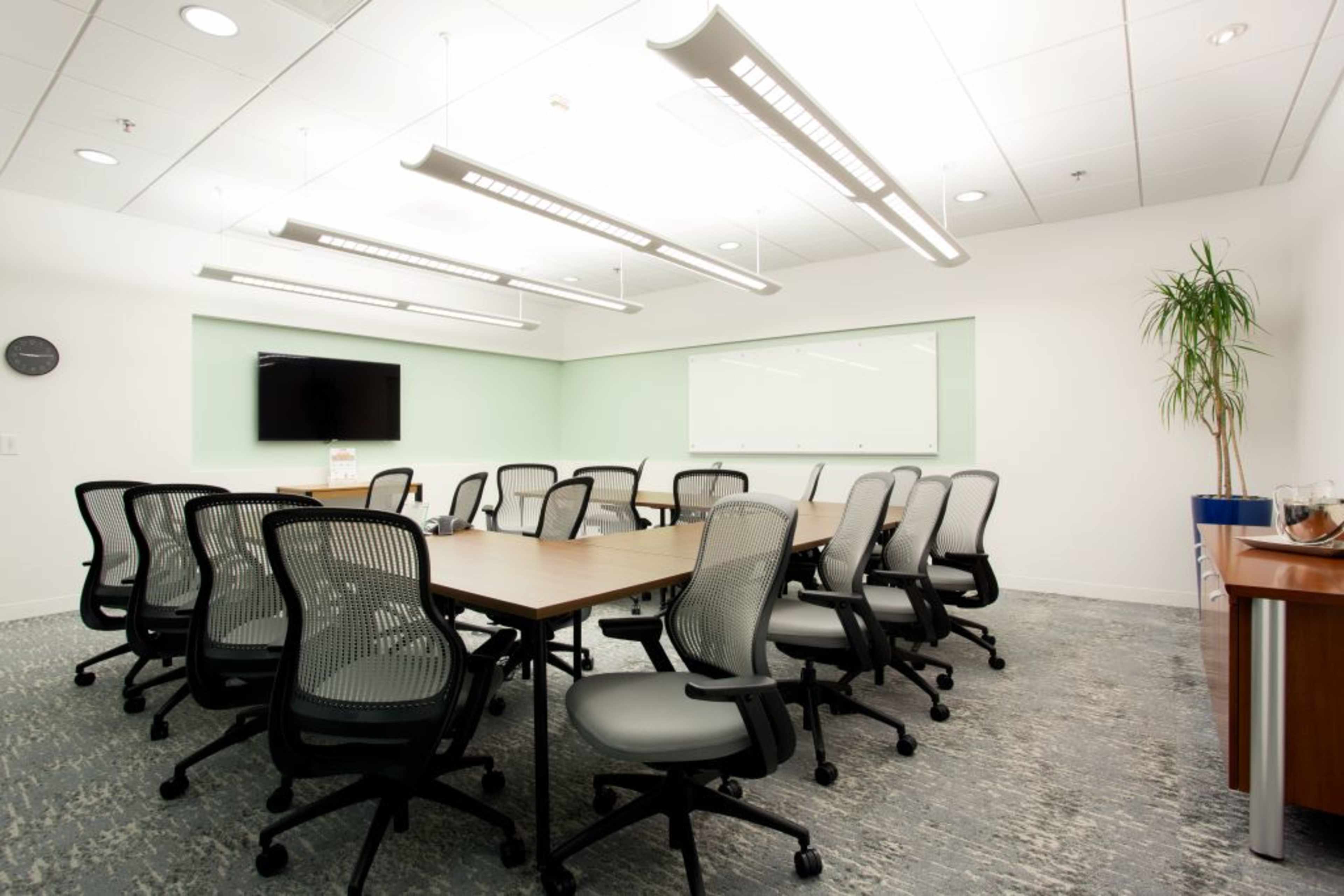 A conference room features a long table surrounded by rolling chairs, with a large screen on one wall and a whiteboard on another.