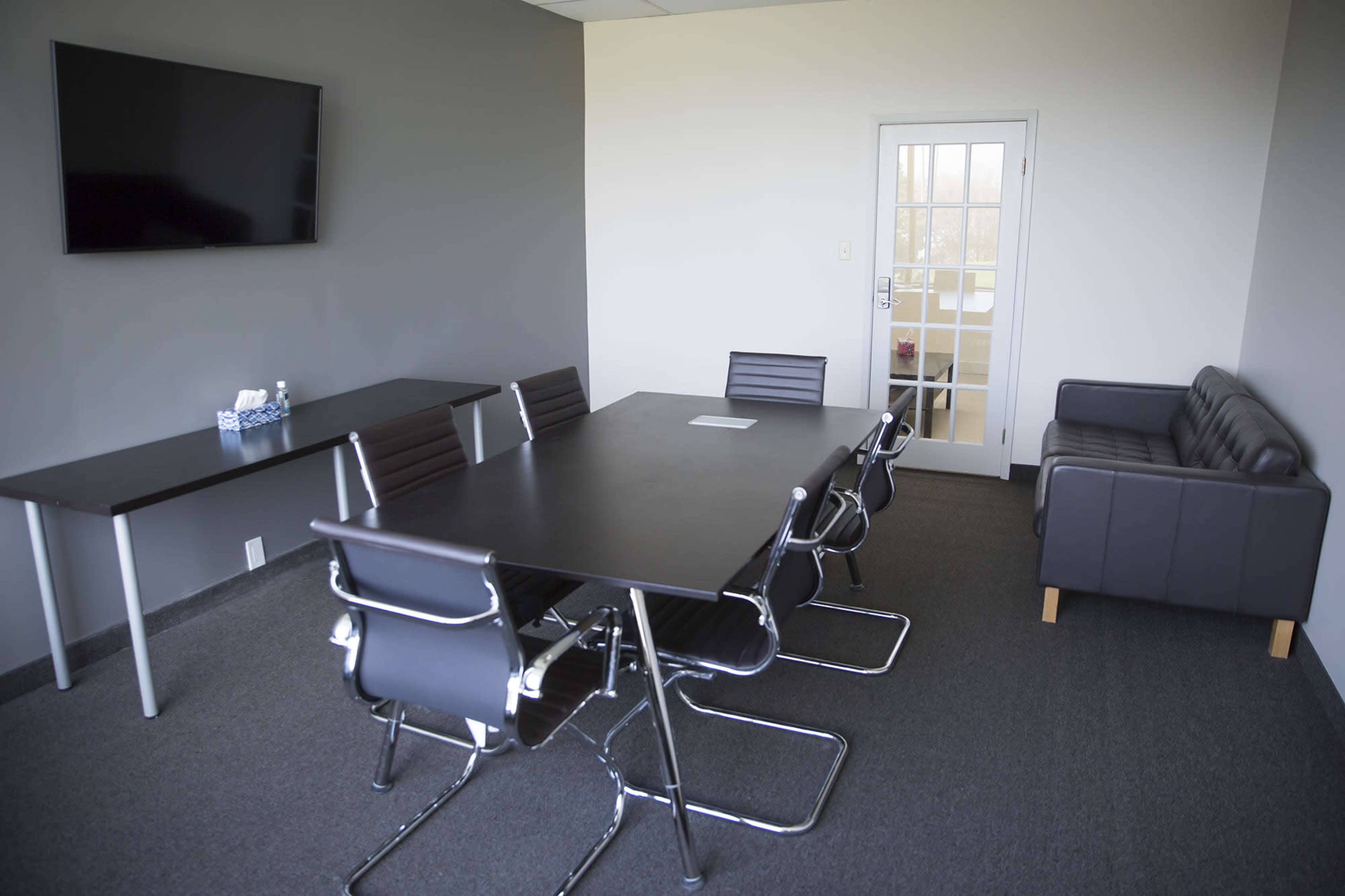 A modern conference room with a long table, several chairs, a television mounted on the wall, and a door leading to another area.