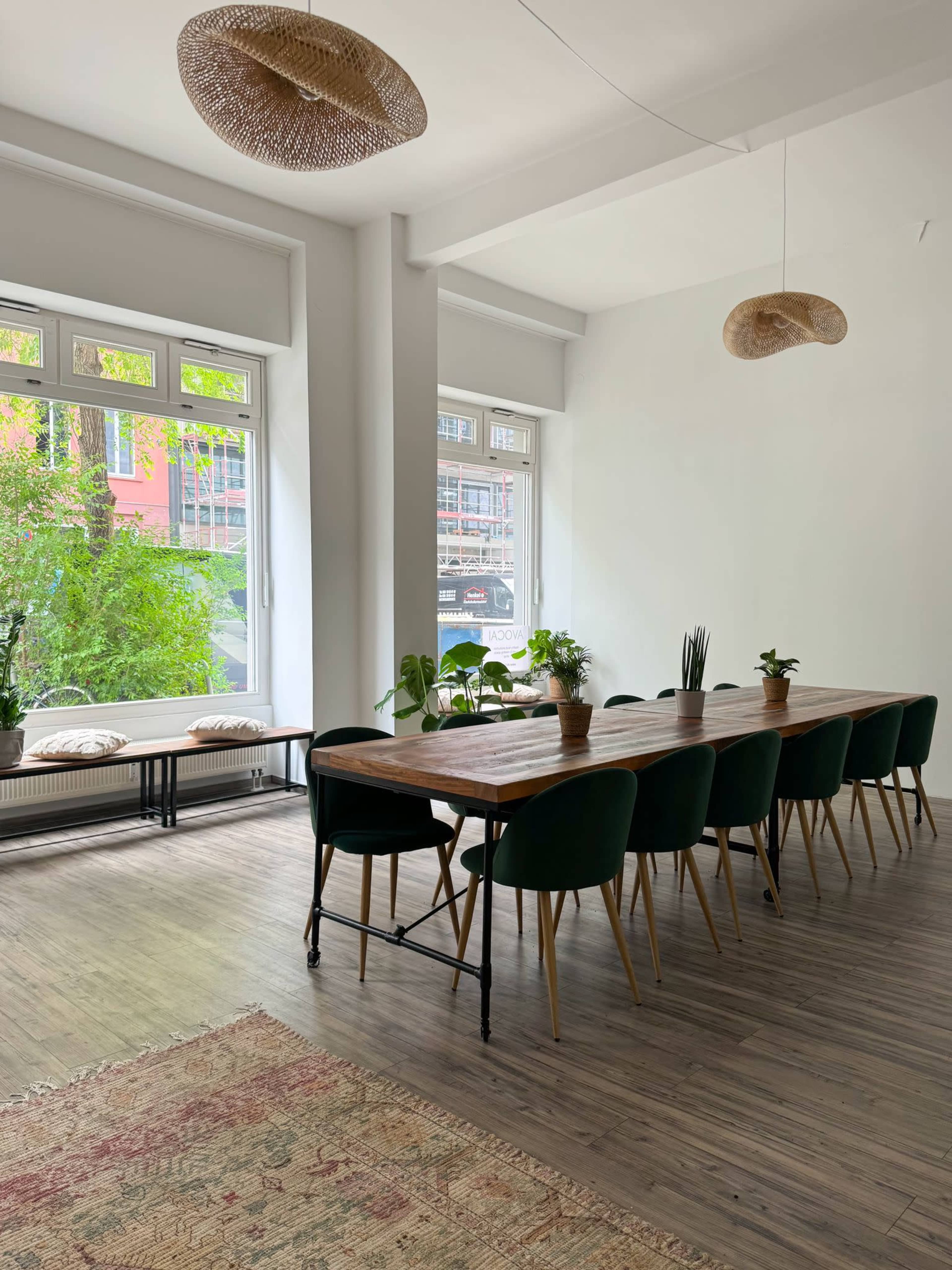 A long wooden dining table with green chairs is set in a bright room featuring large windows and potted plants.