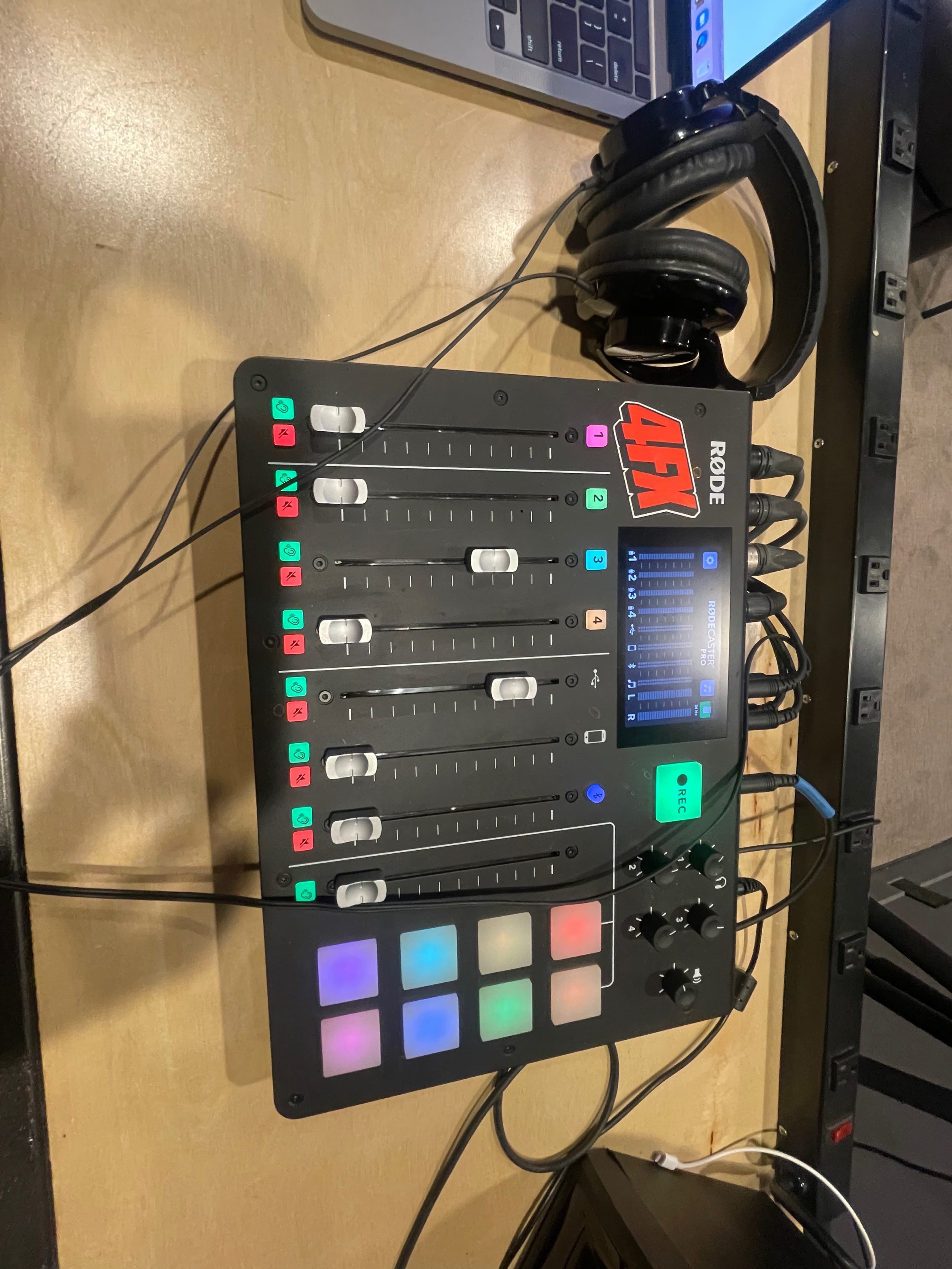 A Rode audio mixer with multiple faders, colored buttons, and headphones resting nearby on a table.