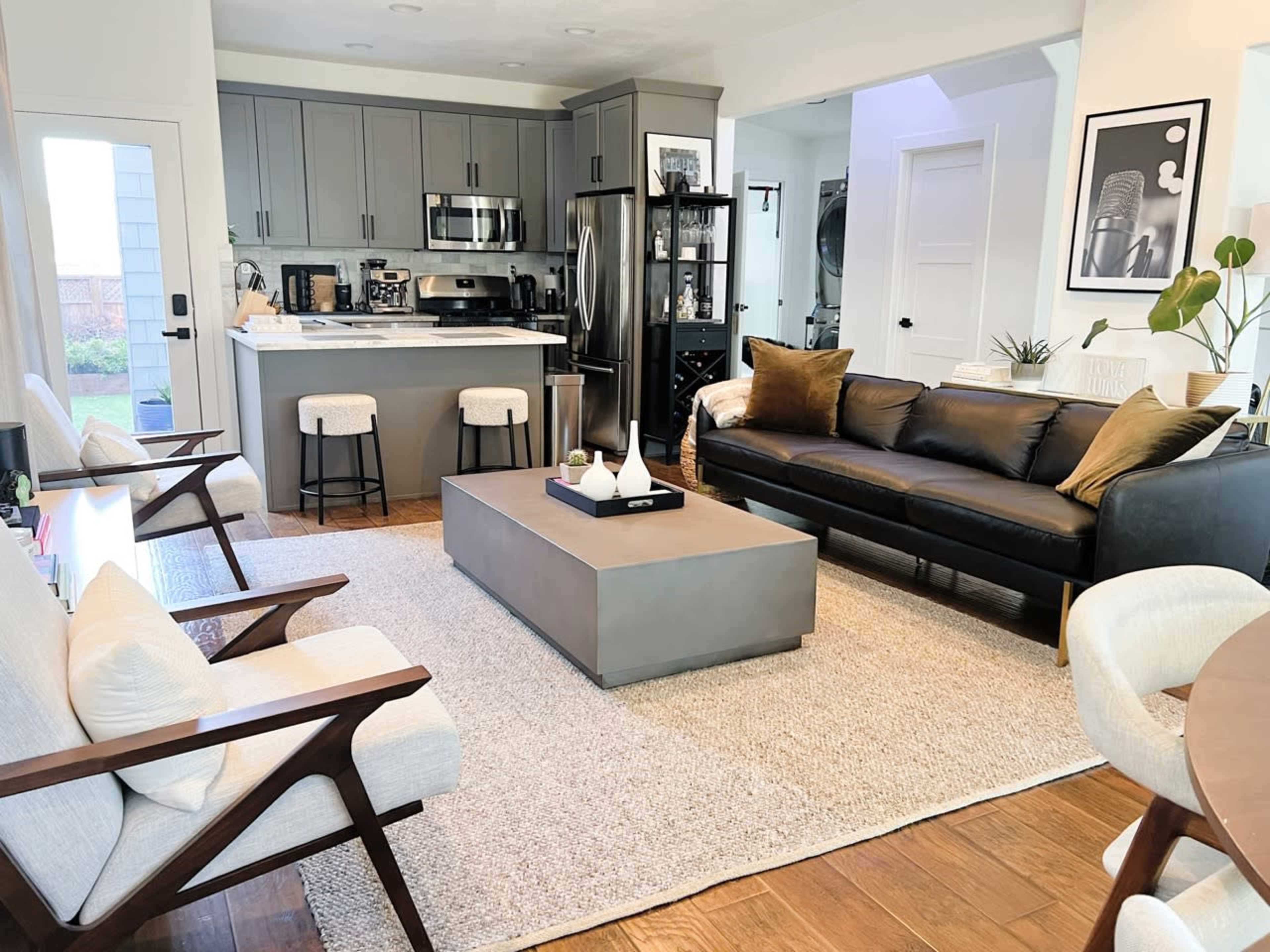 A modern living room features a black leather sofa, a gray coffee table, and a kitchen area in the background with gray cabinets and bar stools.