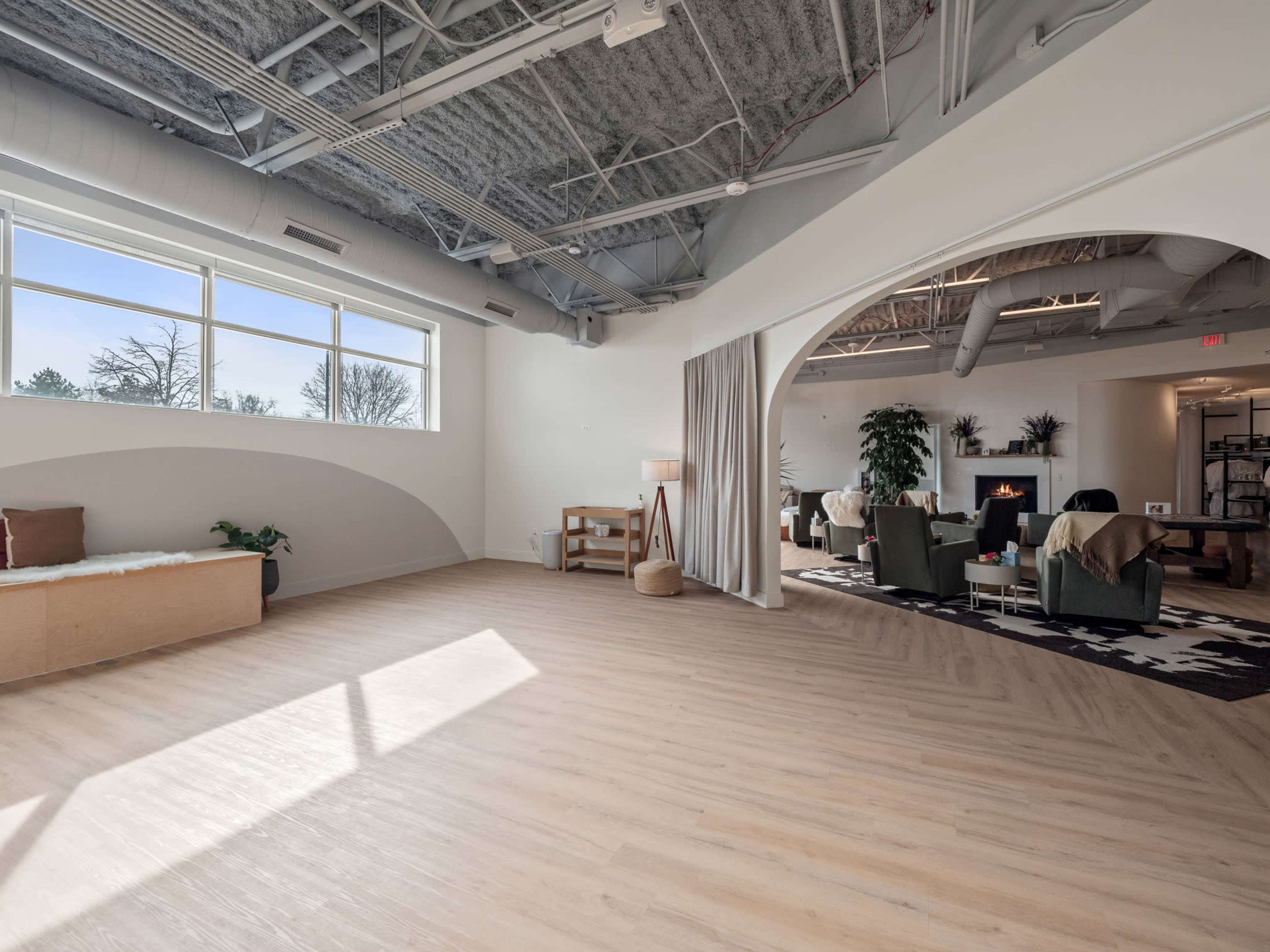 A spacious, well-lit interior with wooden flooring, large windows, and various seating arrangements including a sofa and armchairs.