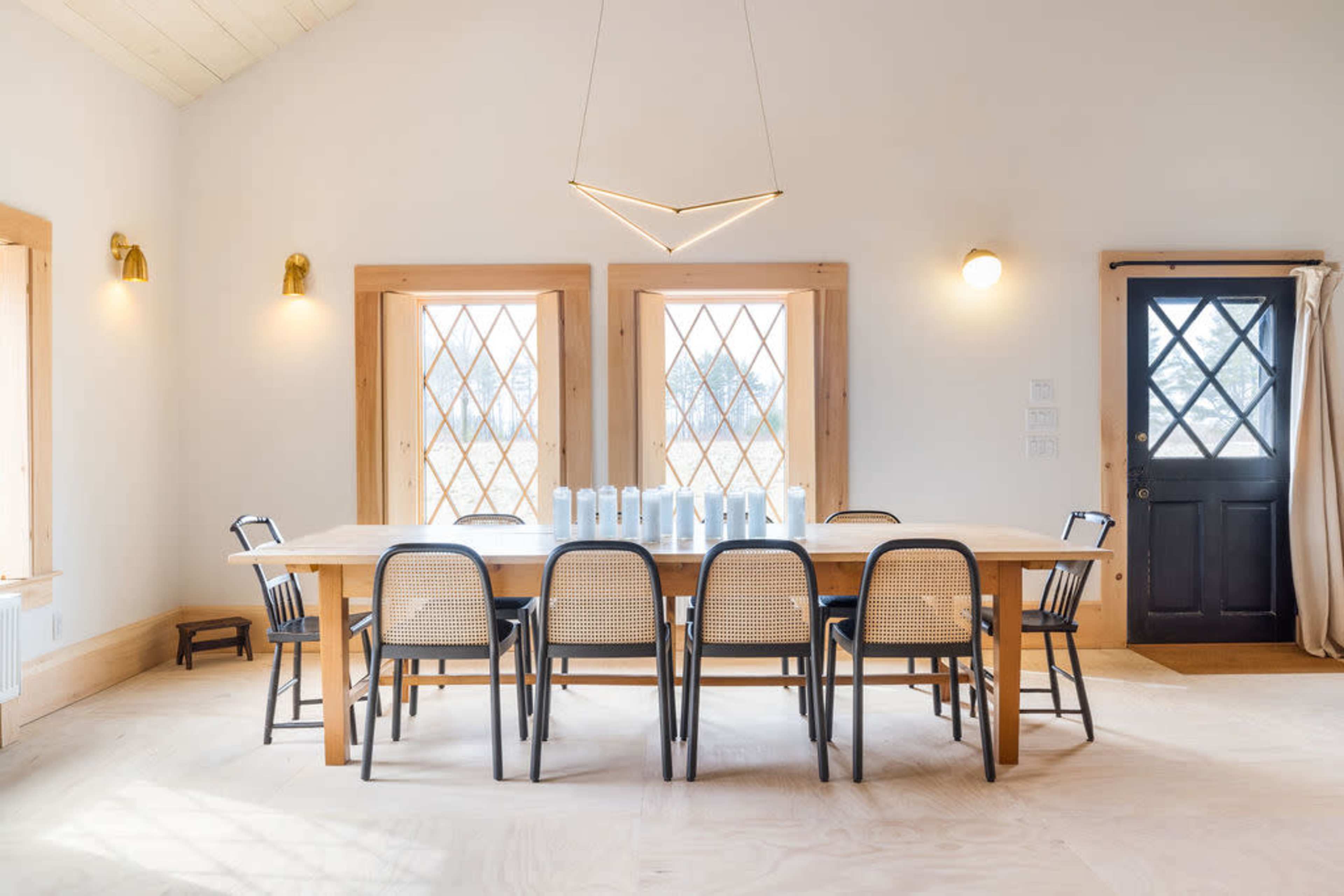 A modern dining room features a long wooden table surrounded by black and cane chairs, with large diamond-patterned windows and a black door.
