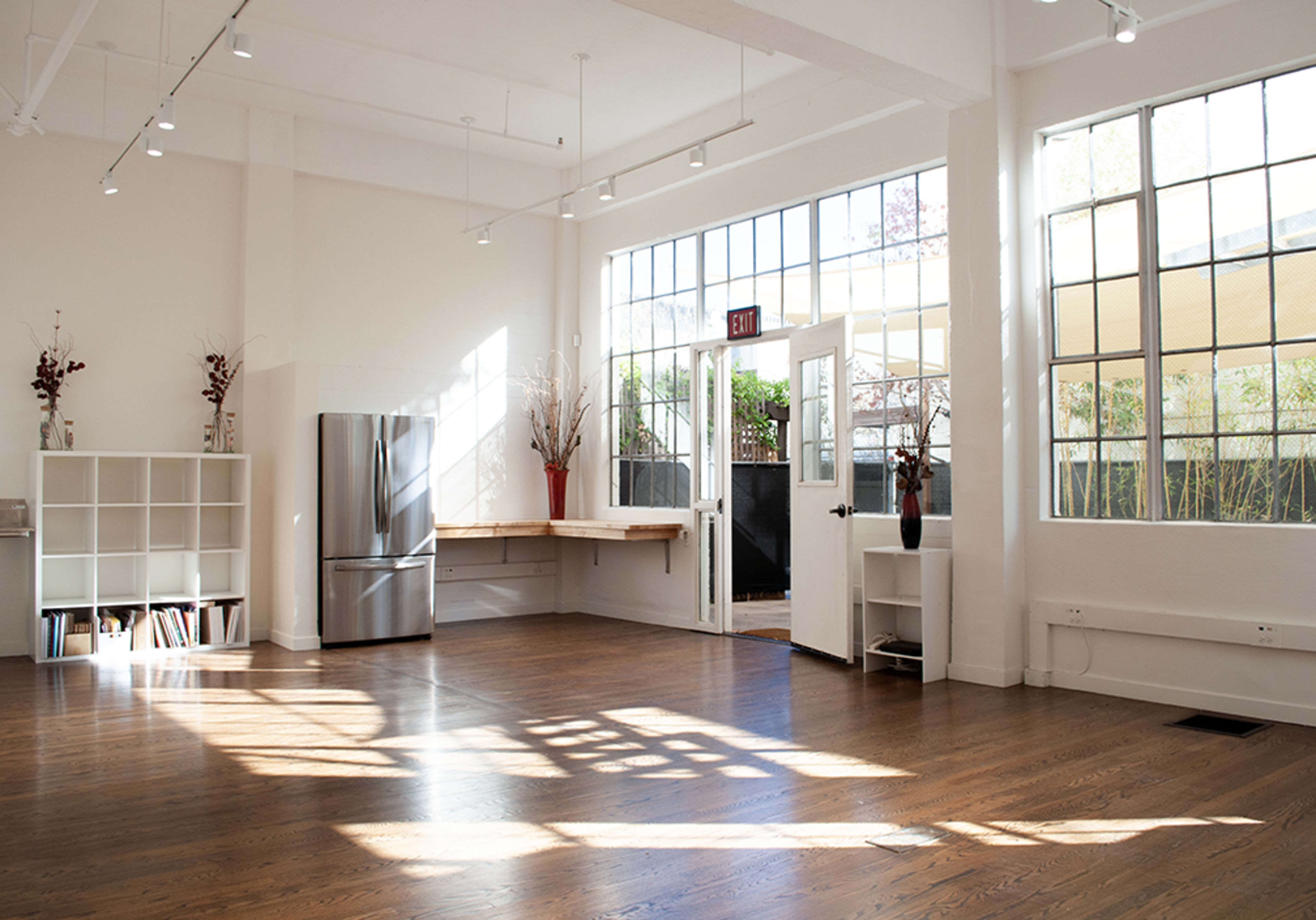 A bright, open interior space with large windows, wooden floors, and minimalist furniture, including a stainless steel refrigerator and a shelf with books.