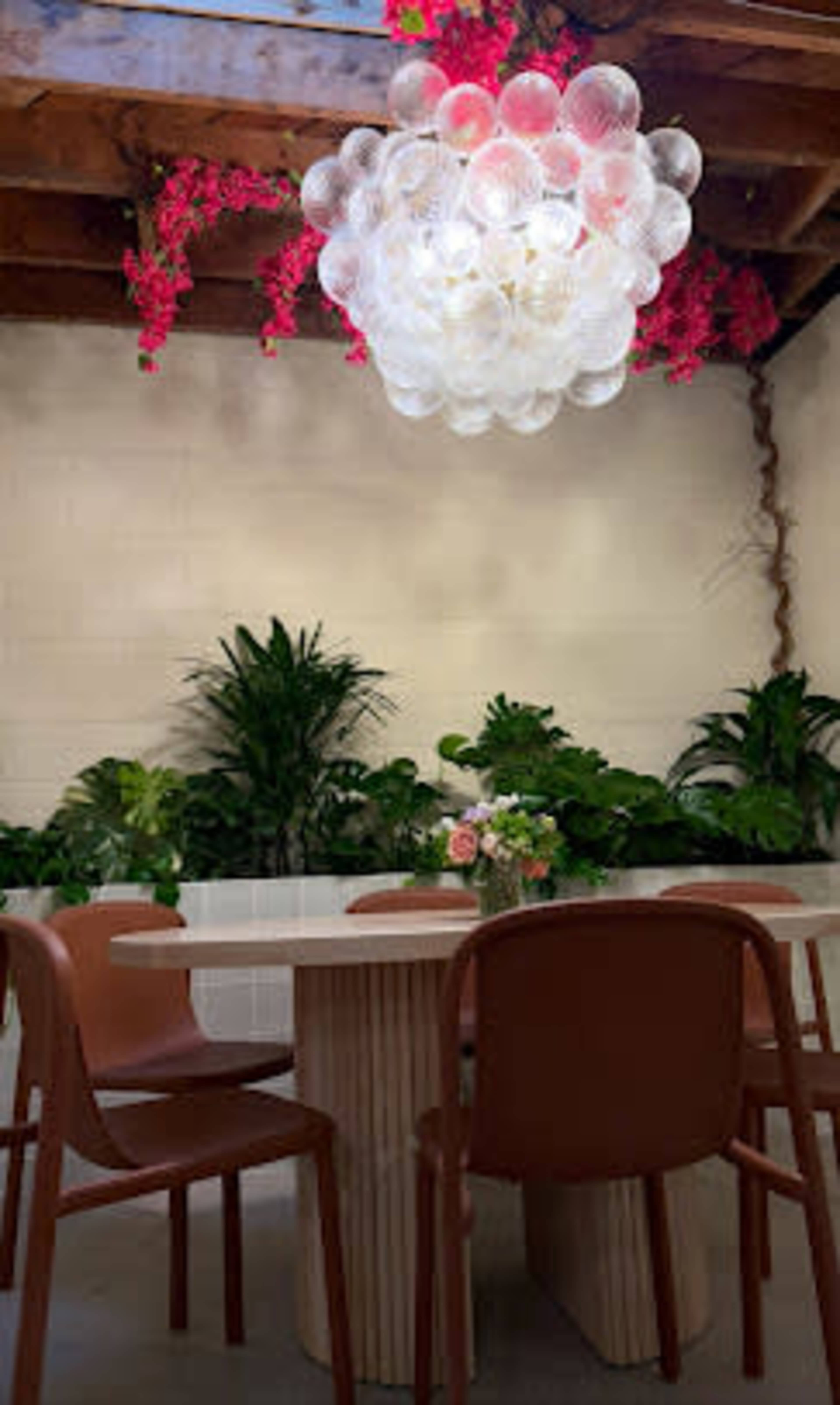 A dining area features a round table surrounded by brown chairs, with a large bubble chandelier and greenery along the walls.