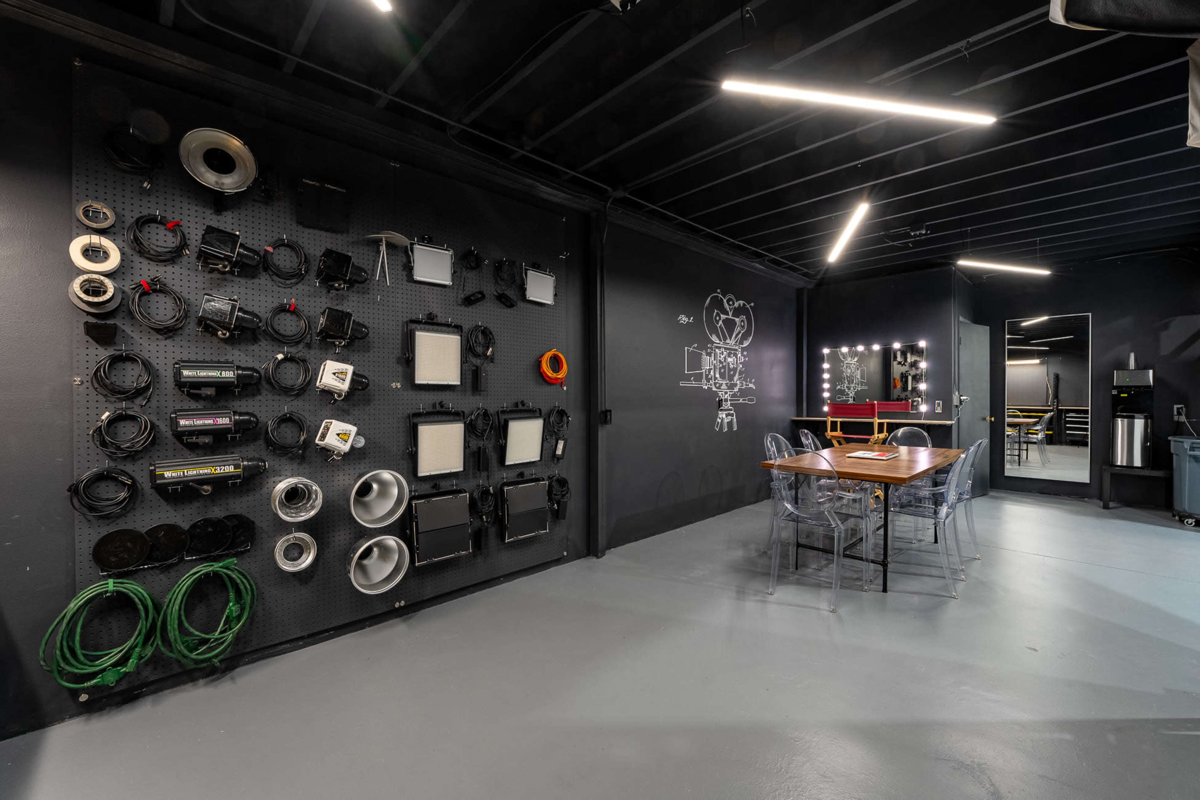 A modern interior space featuring a wall with various audio-visual equipment and cables, a table with clear chairs, and a well-lit area with mirrors.