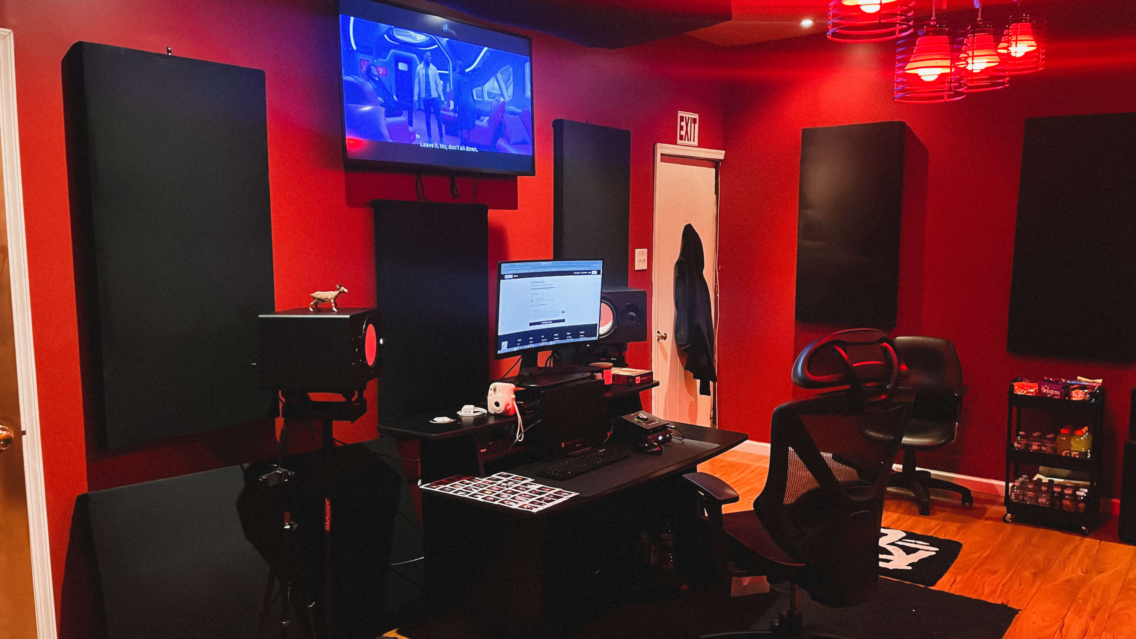 The image shows a recording studio featuring a computer desk with monitors, a red-painted wall, acoustic panels, and a television mounted on the wall.