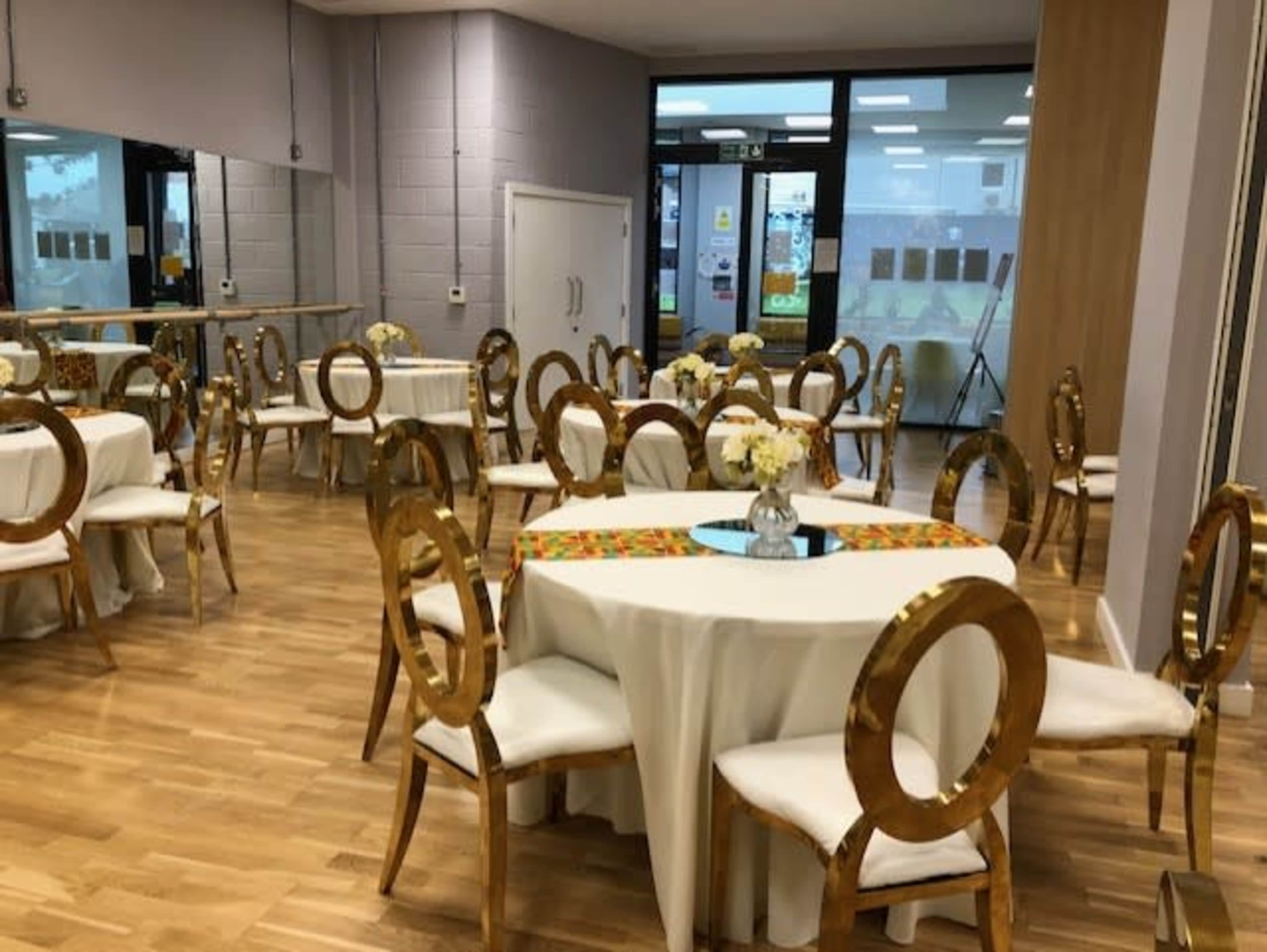 A spacious indoor venue features several round tables with white tablecloths, gold chairs, and floral centerpieces.