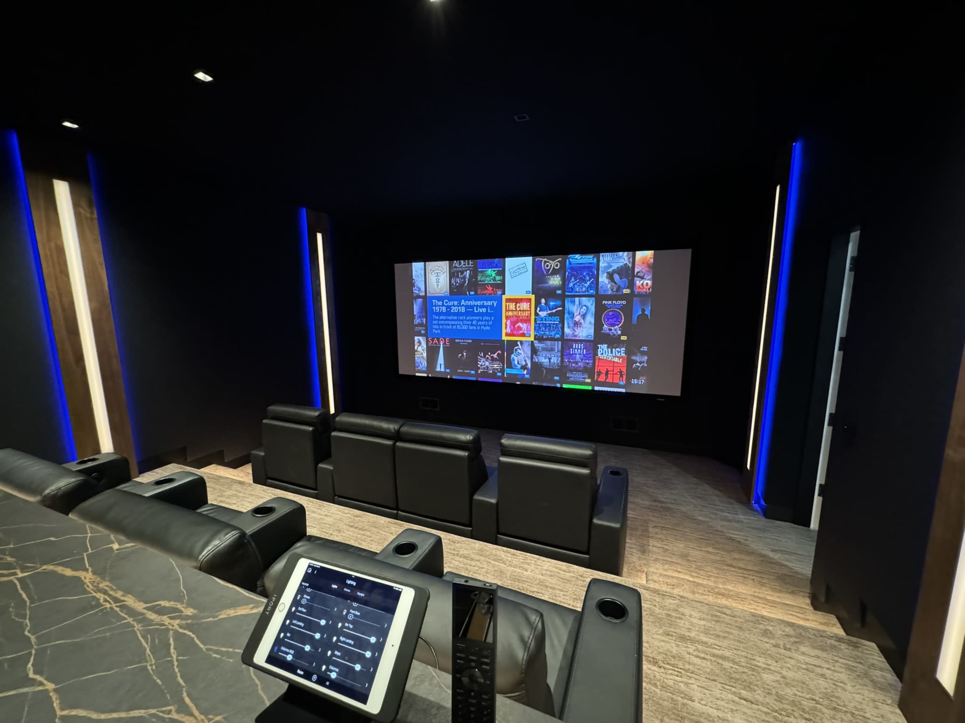 A dark home theater featuring a large screen displaying a media library, with black reclining chairs arranged in front.
