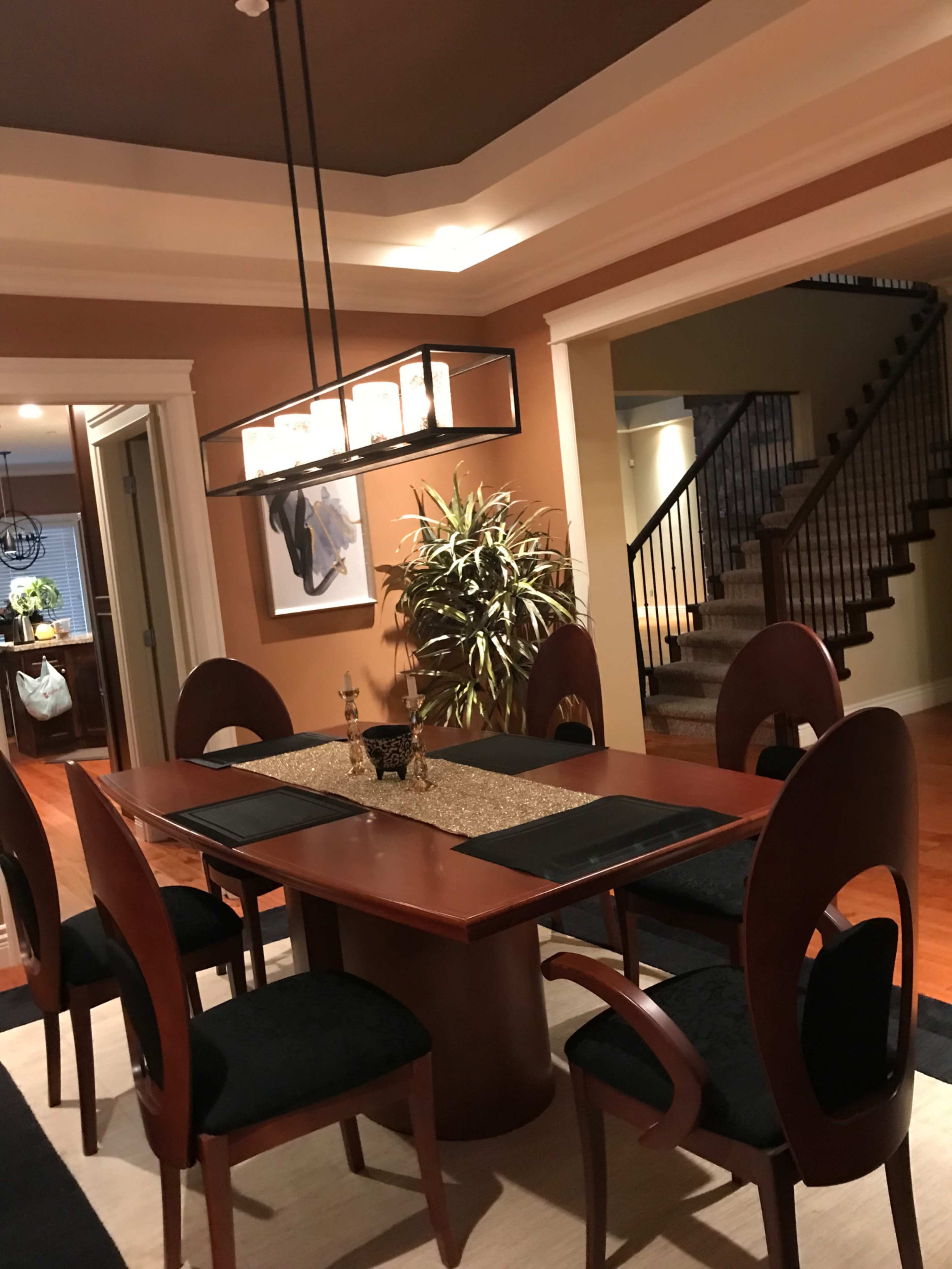A dining area with a rectangular wooden table surrounded by black chairs, a pendant light above, and a staircase leading to another level in the background.