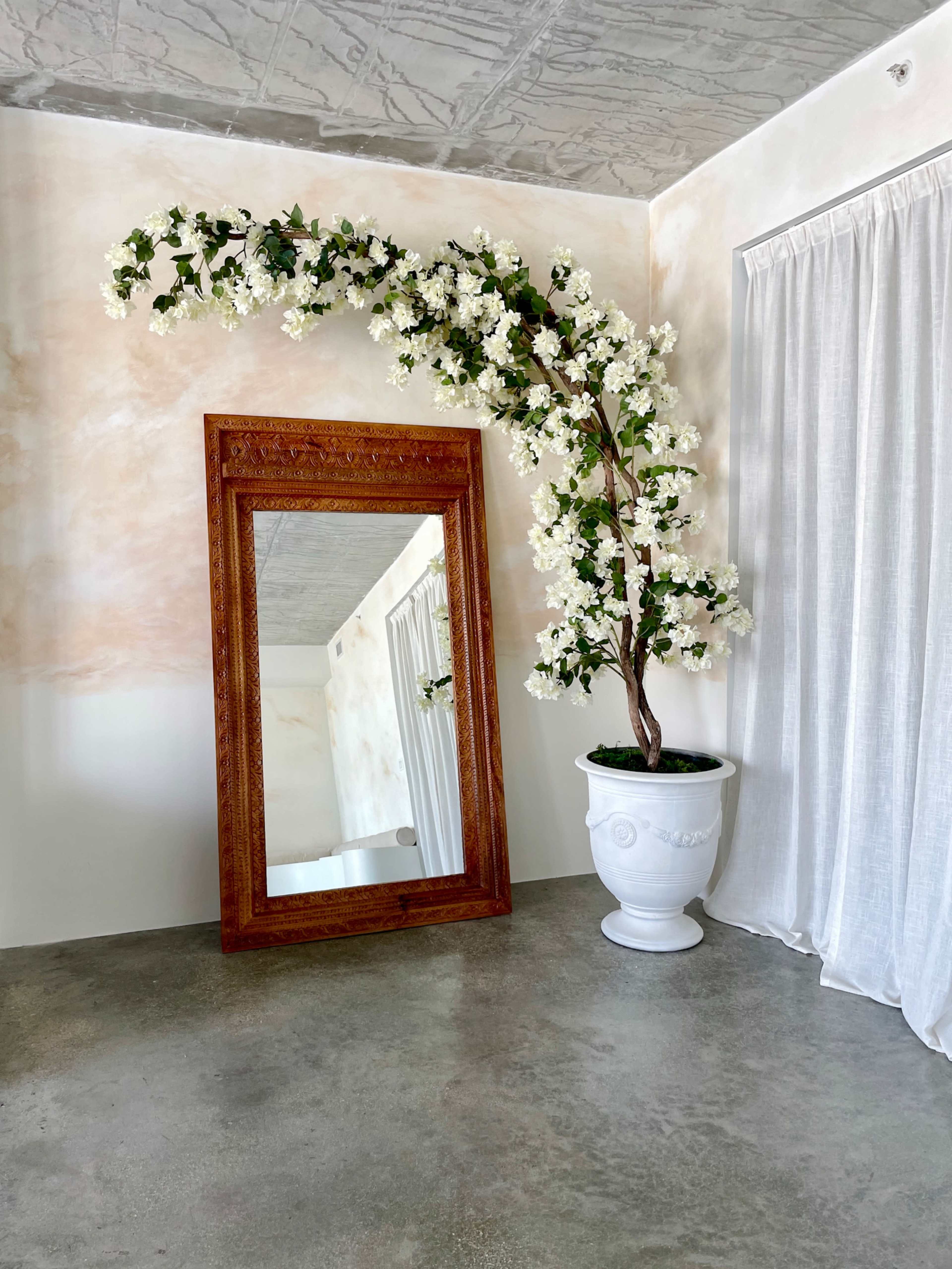 The image shows a large ornate mirror reflecting a decorative branch of white flowers in a pot, set against a softly colored wall and draped curtains.