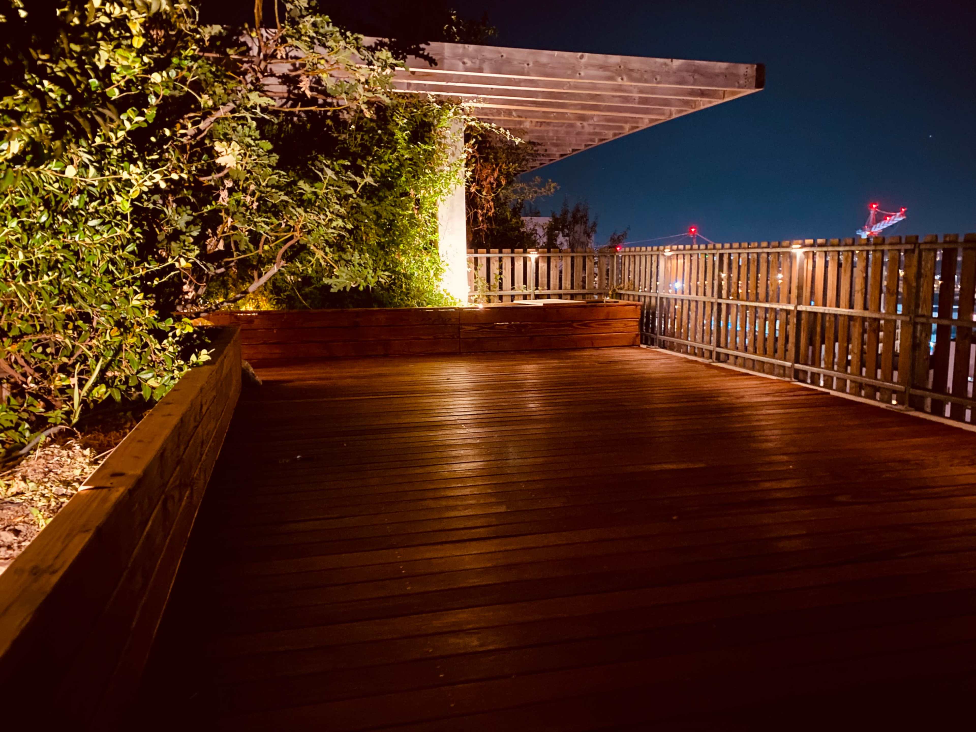 The image shows a wooden deck surrounded by greenery and a partially covered area, illuminated at night.