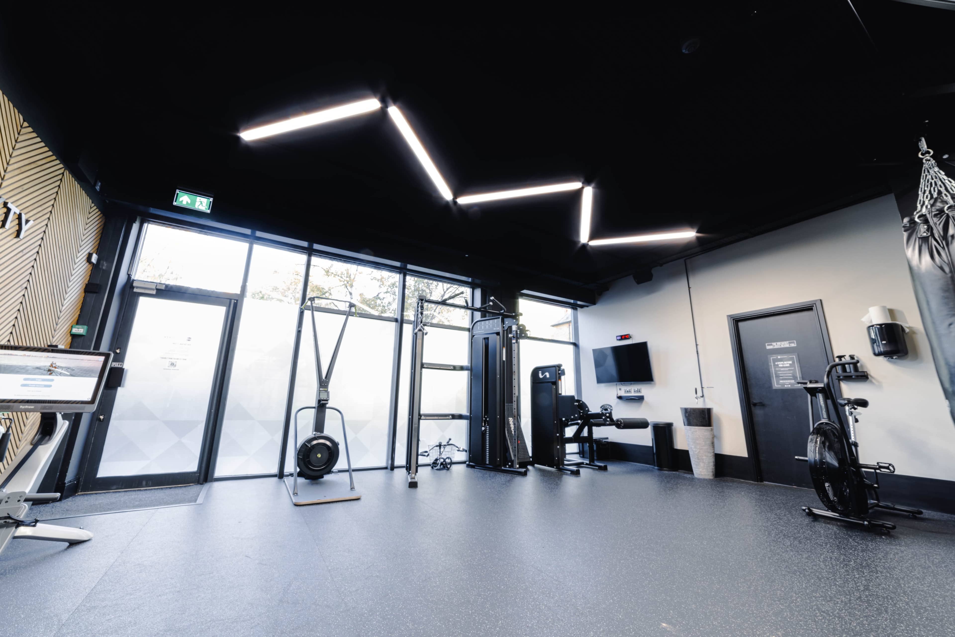 The image shows a modern gym interior featuring various exercise equipment, large windows, and a ceiling with unique lighting design.