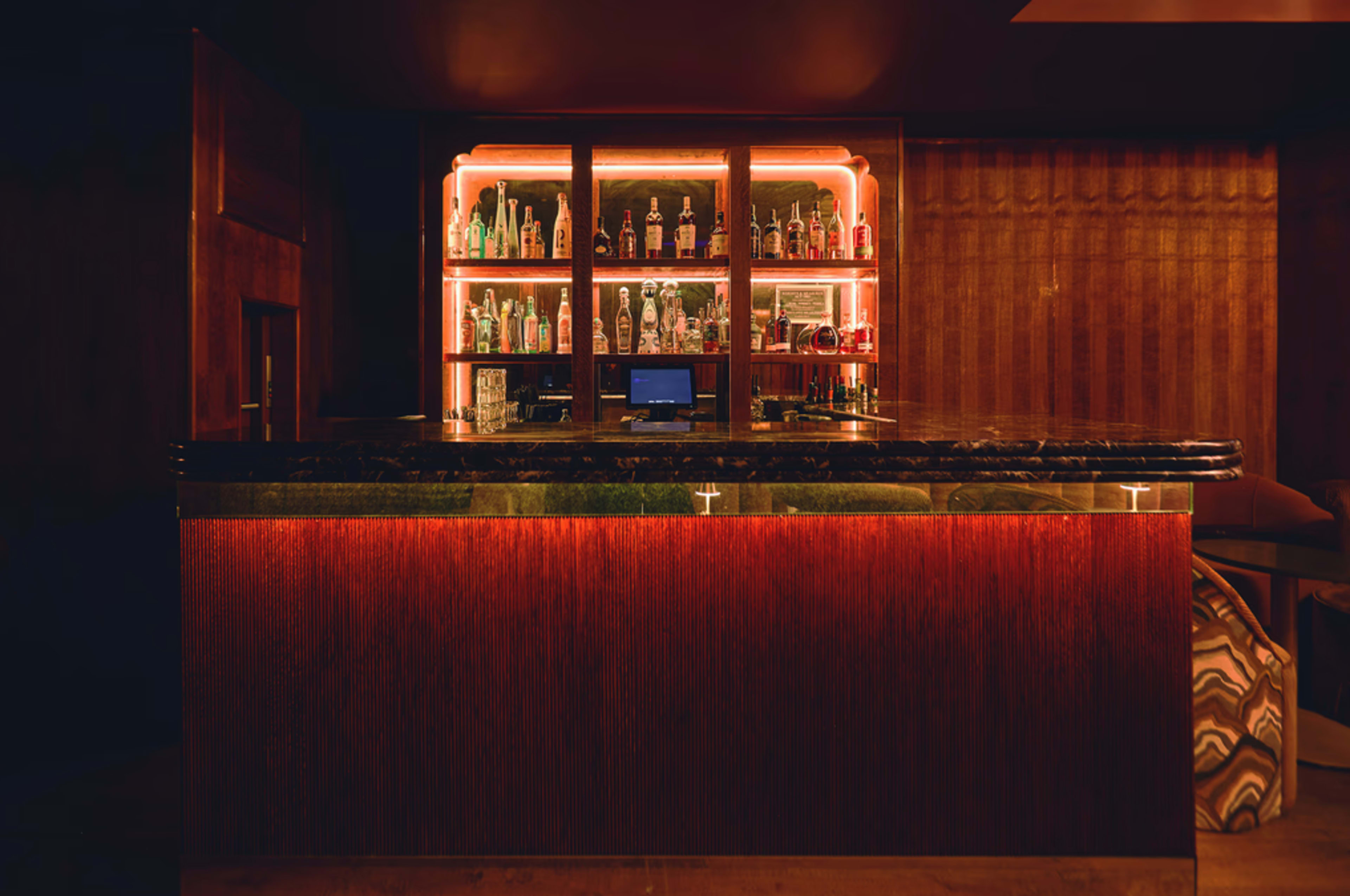 The image shows a dimly lit bar with a sleek countertop and a display of various bottles illuminated by soft lighting in the background.