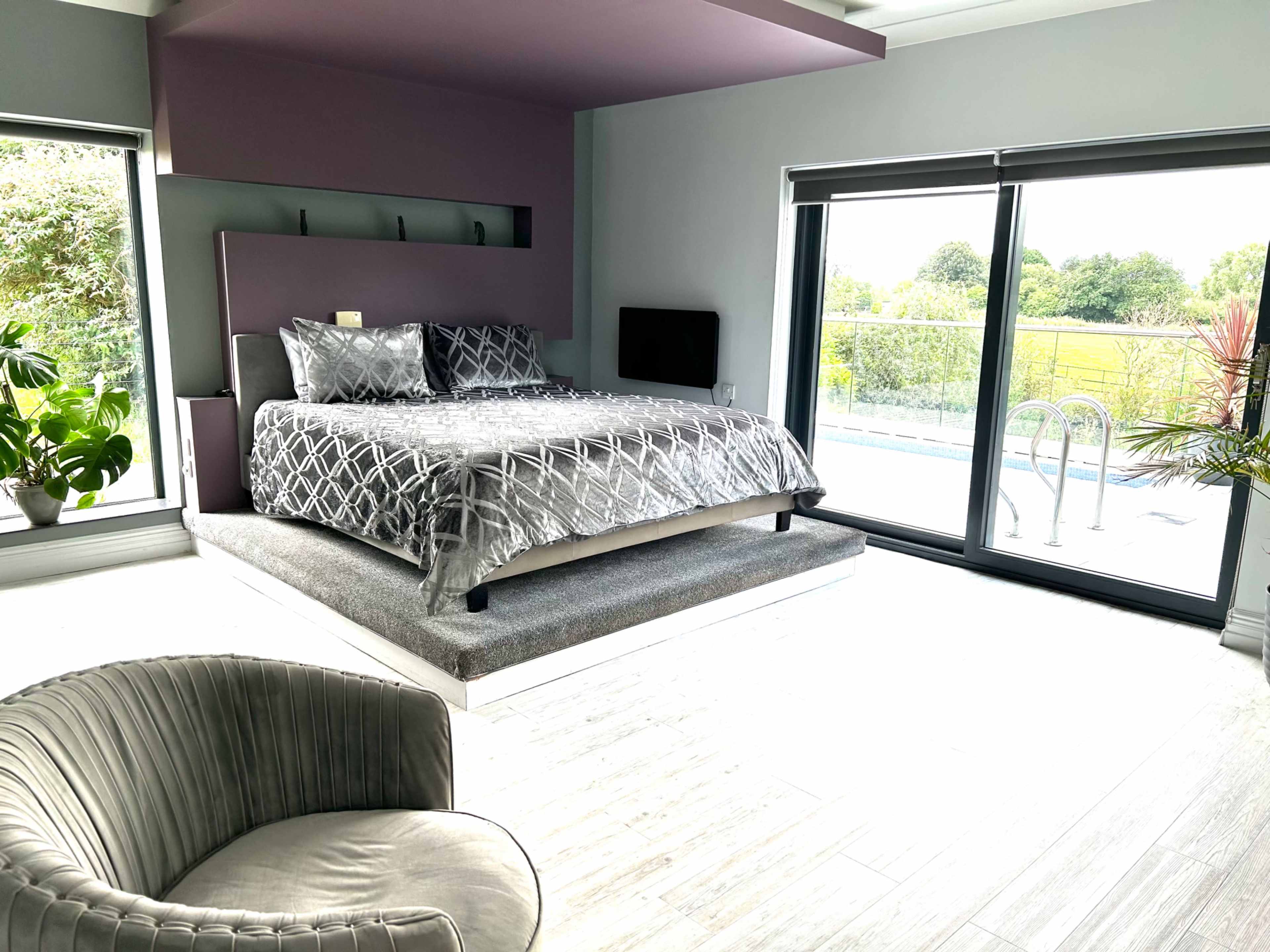 A modern bedroom features a raised bed with decorative pillows, a flat-screen TV, and large sliding glass doors leading to an outdoor area.