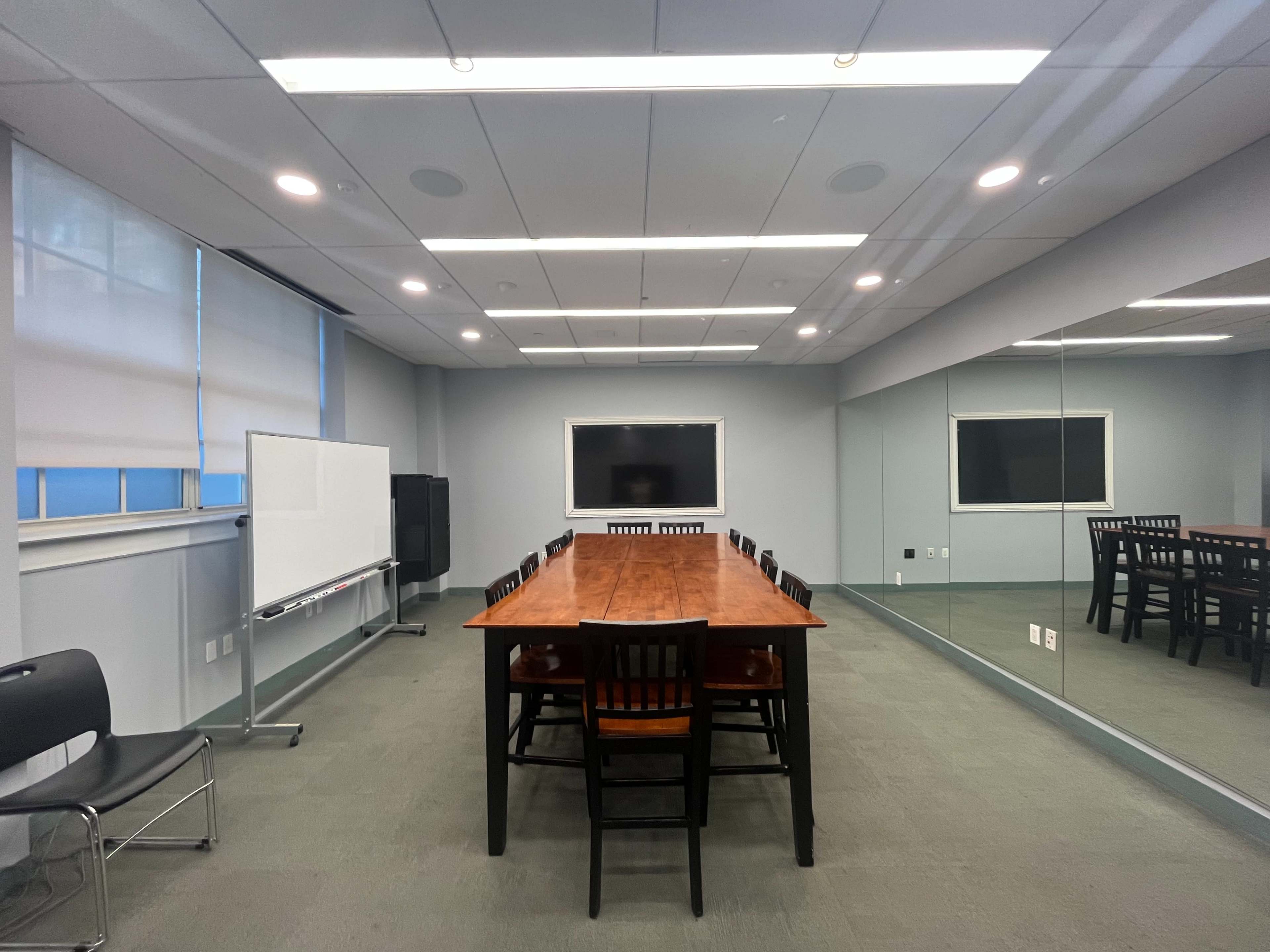 The image depicts a spacious conference room with a long wooden table surrounded by black chairs, a whiteboard, and a television mounted on the wall.