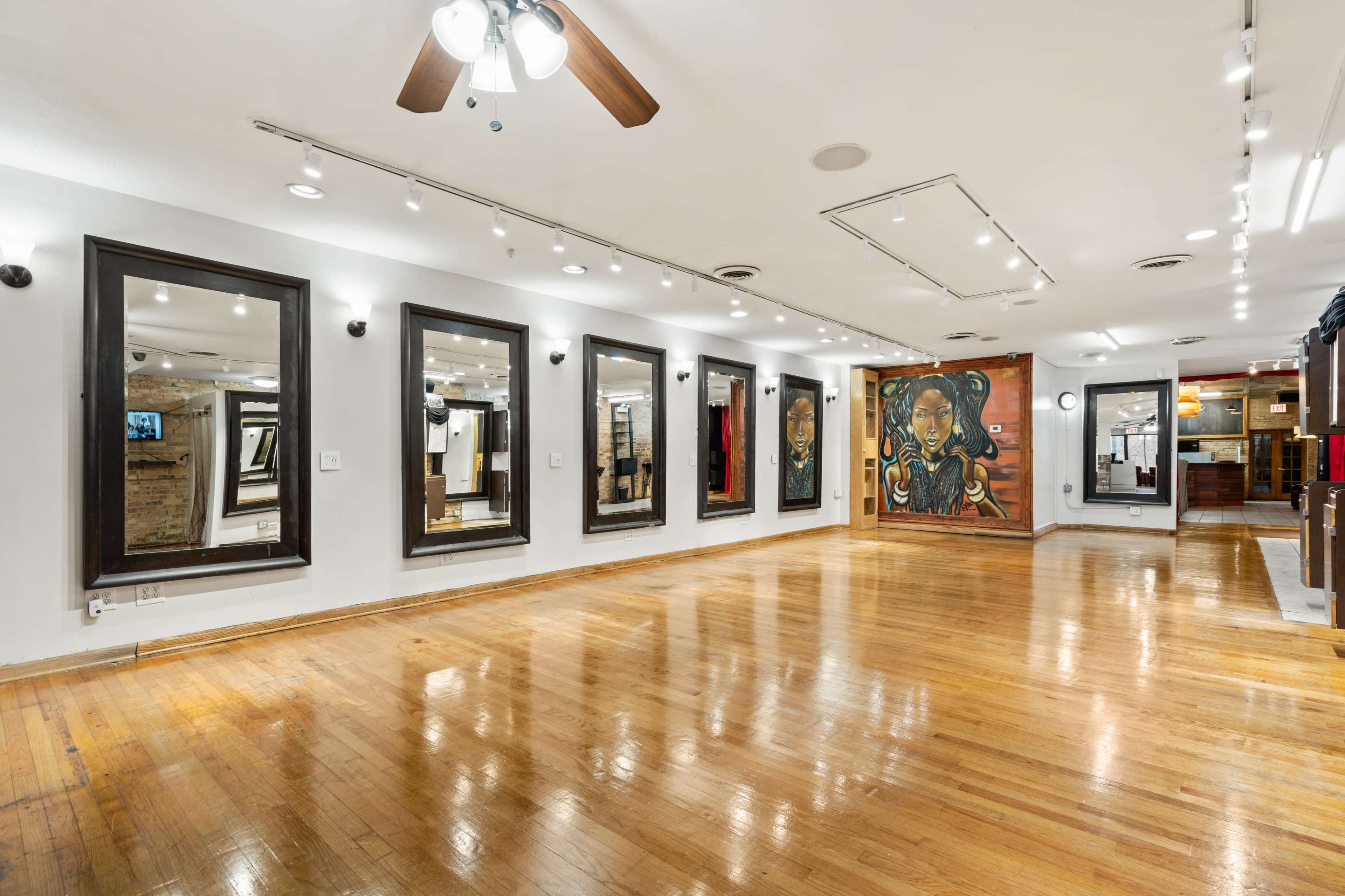 The image shows a spacious gallery with polished wooden floors, featuring several large mirrors on the walls and a prominent mural of a woman.