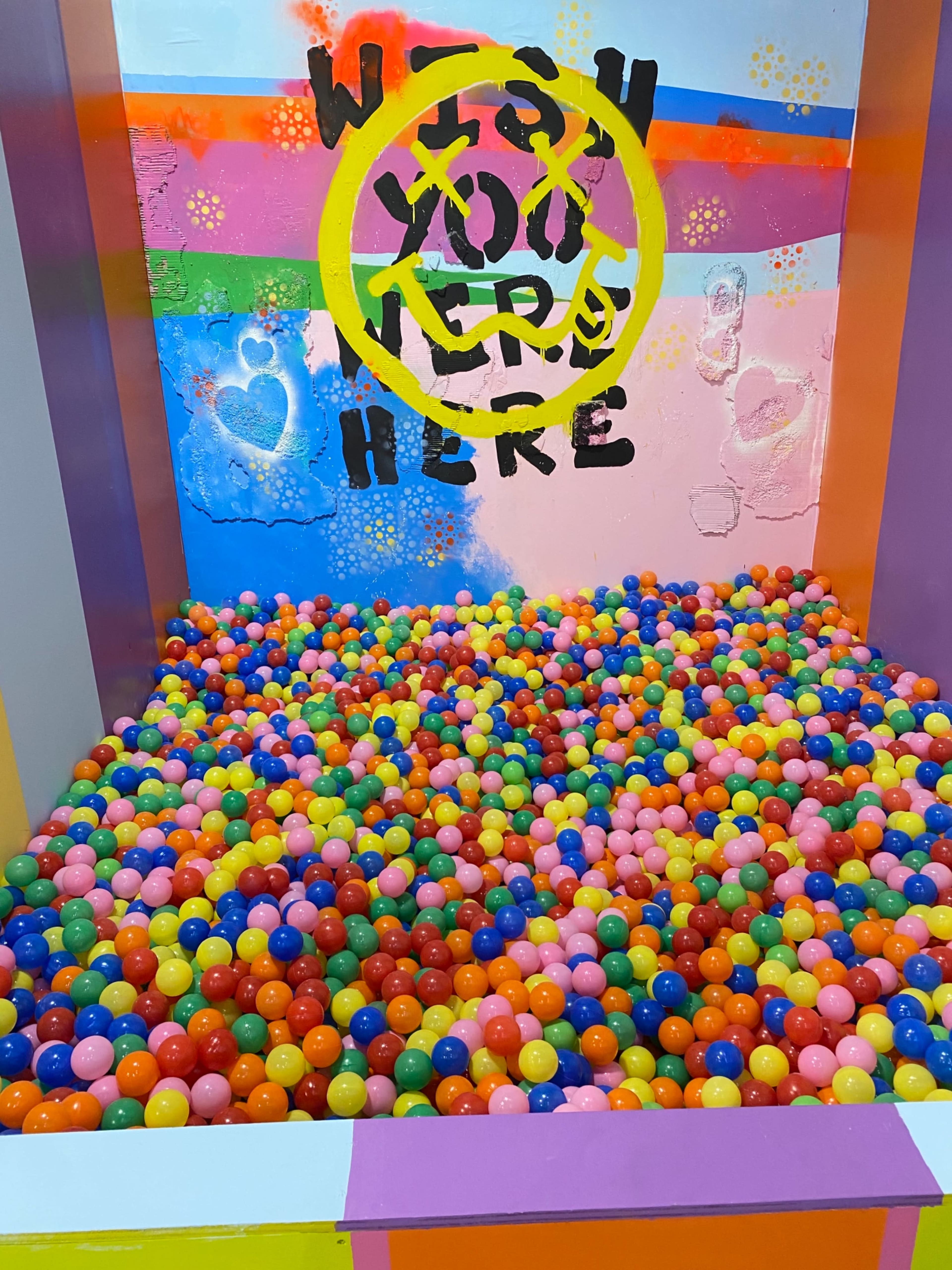 A brightly colored ball pit filled with a variety of plastic balls in multiple hues, set against a backdrop featuring painted messages and designs.