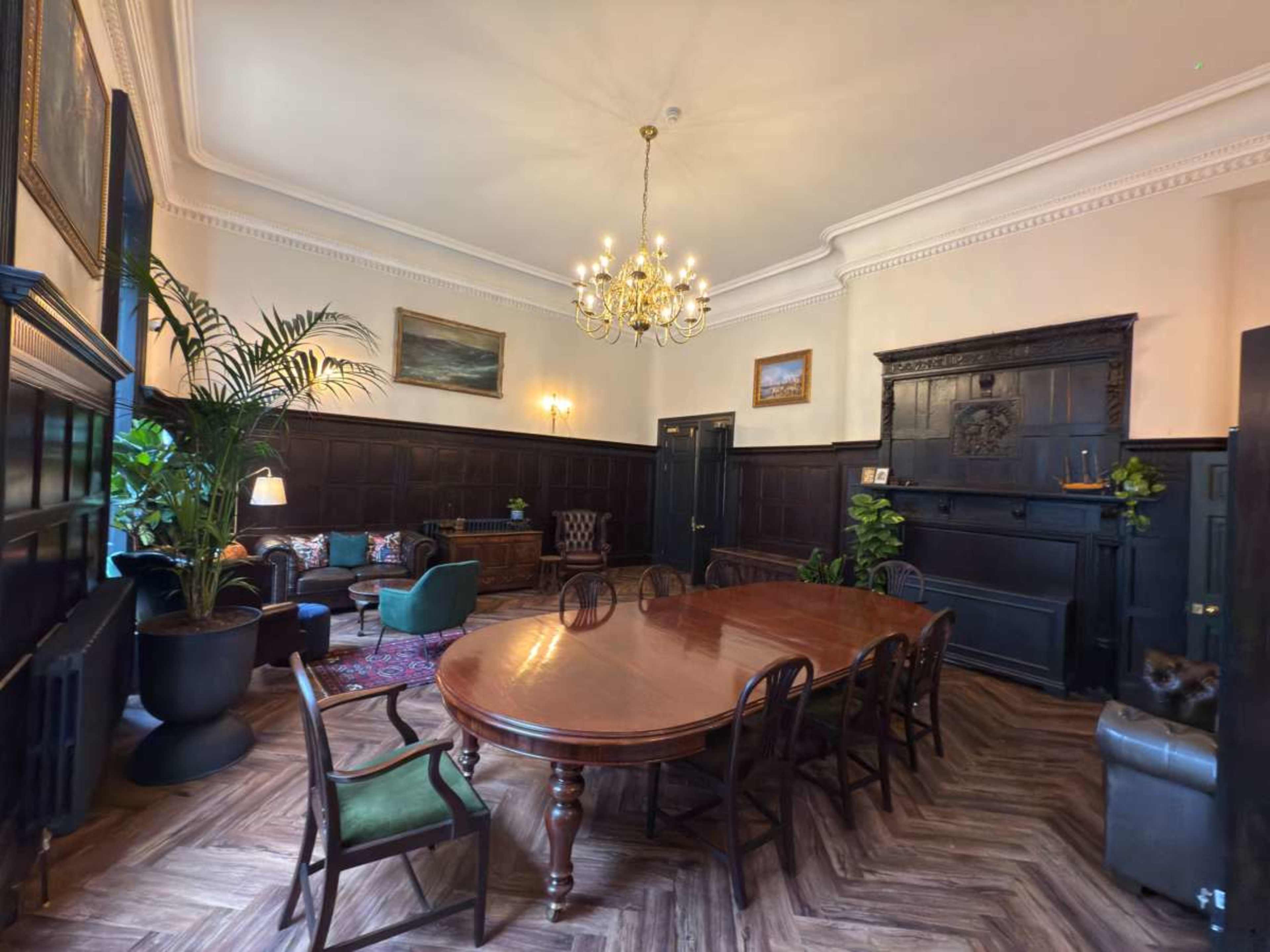 The image depicts a well-furnished room featuring a large wooden dining table surrounded by chairs, complemented by a chandelier and decorative plants.