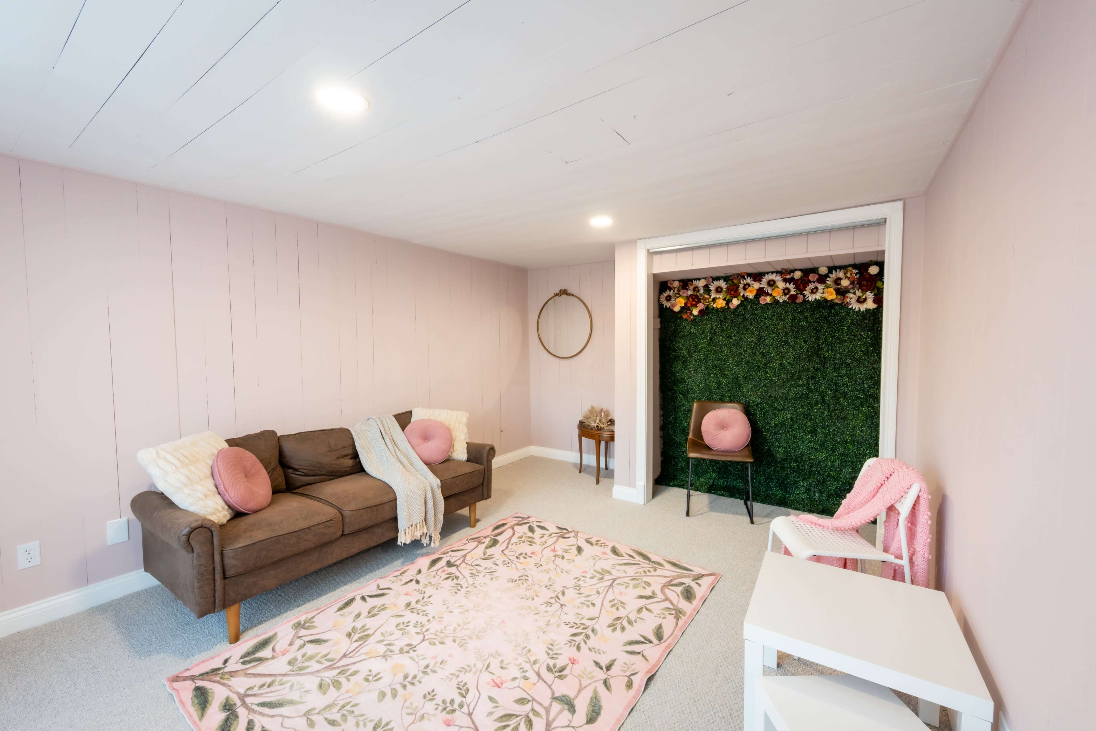 The image shows a cozy, light pink-walled room furnished with a brown sofa, decorative pillows, a floral backdrop, and a plush area rug.