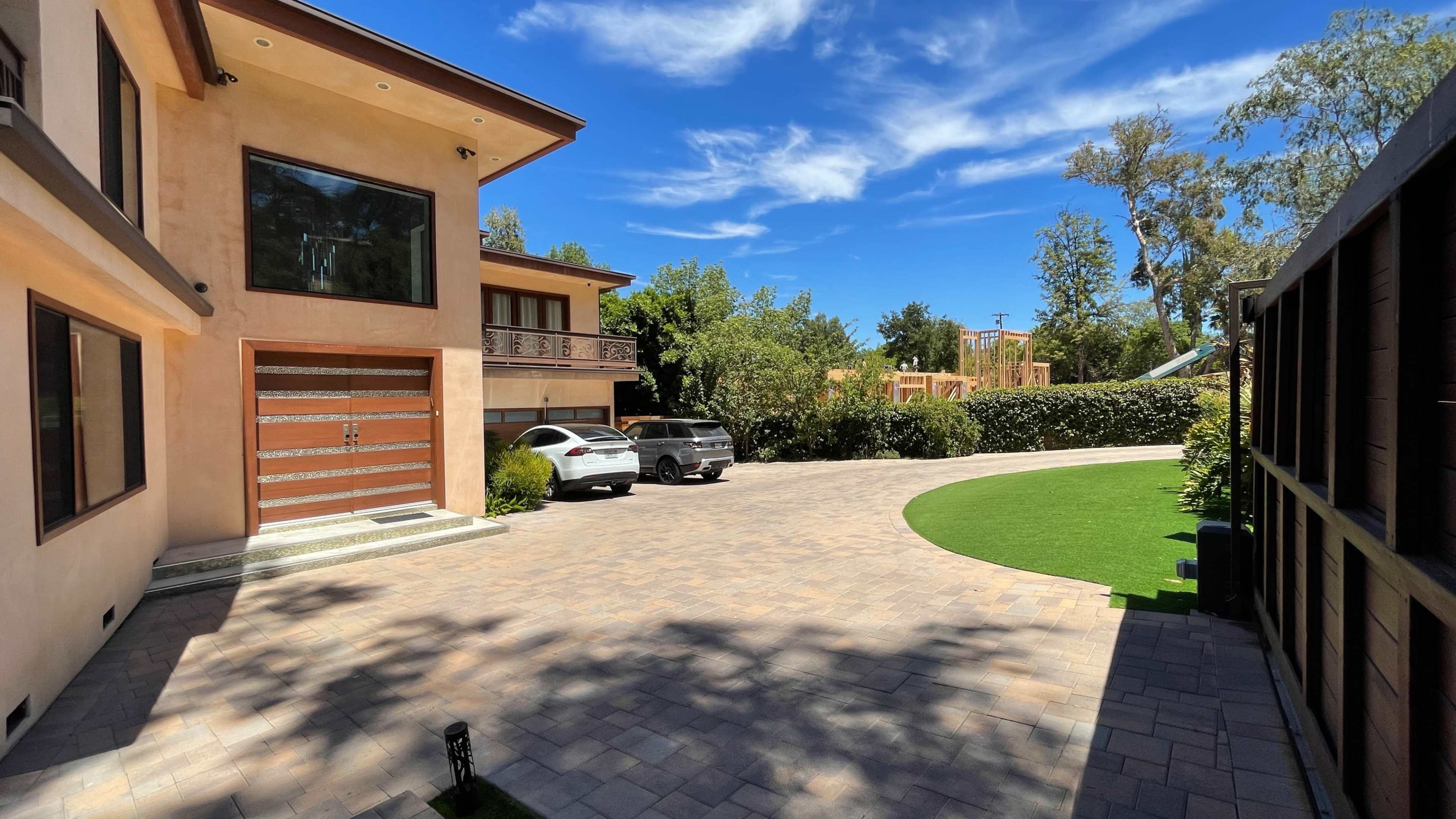 A residential driveway leads to a two-story house with a wooden garage door, and two parked cars are visible against a backdrop of greenery and a clear blue sky.