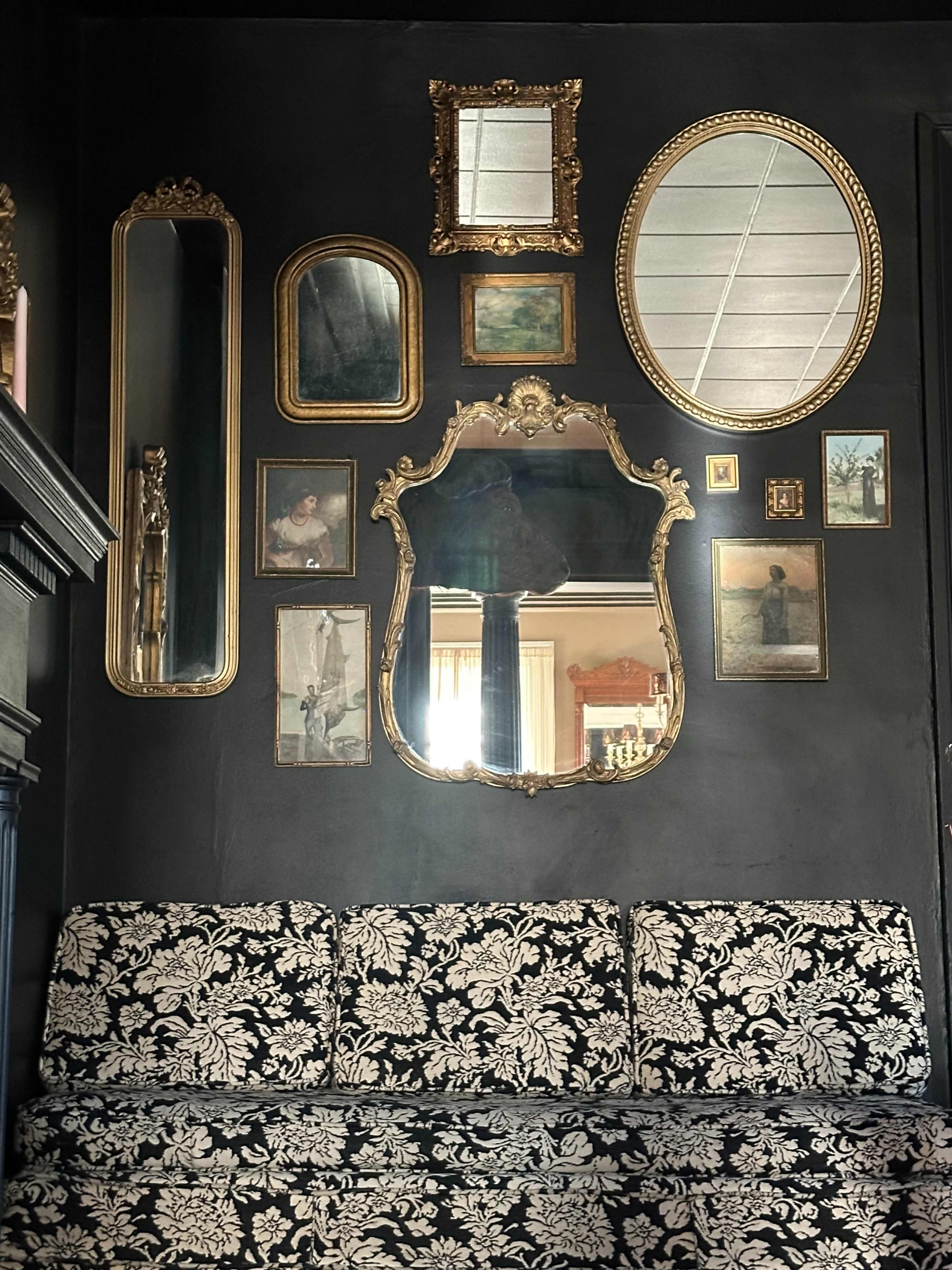 A black wall features a variety of ornate mirrors and framed artwork above a floral-patterned sofa.