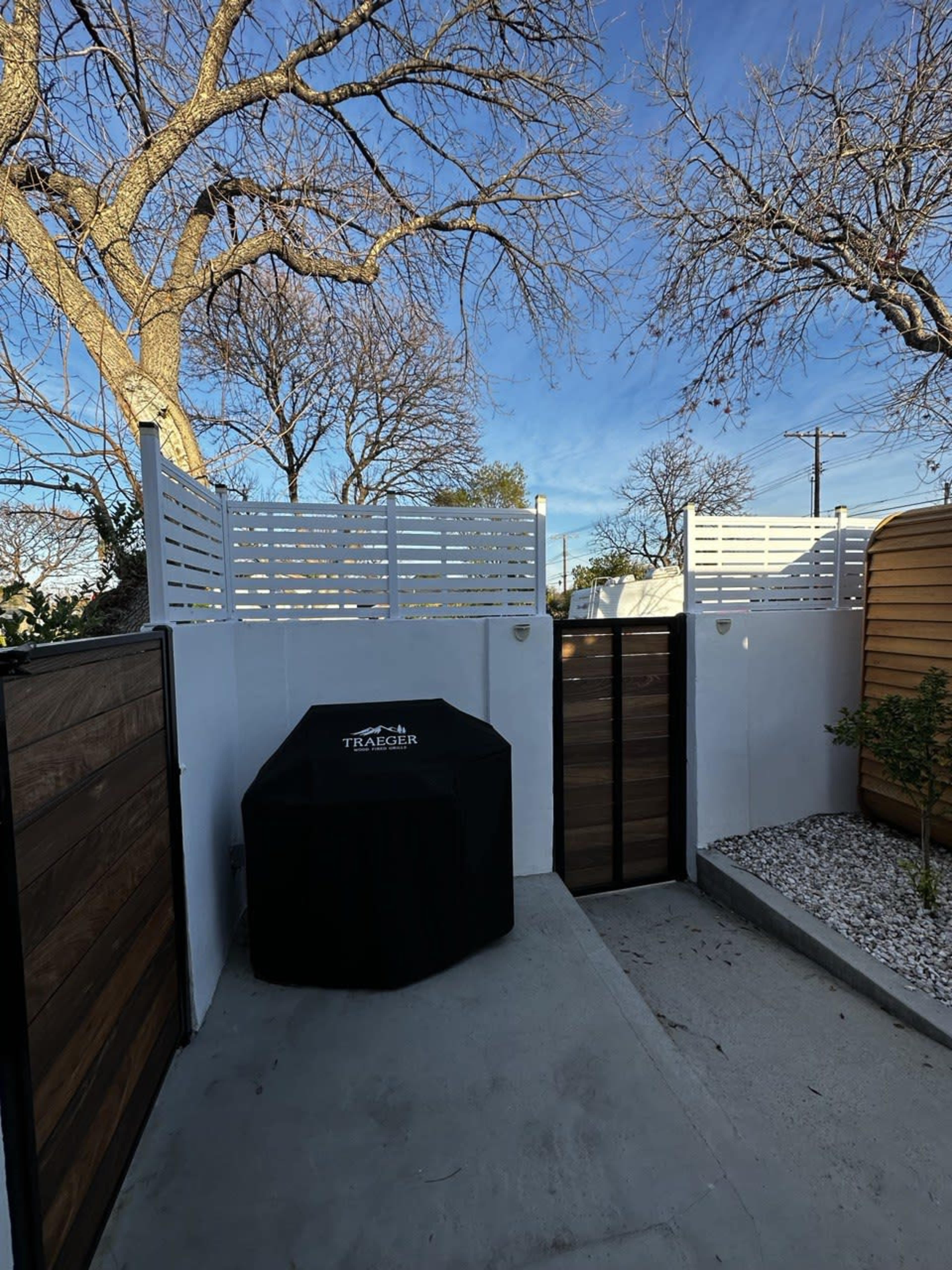 A patio area is enclosed by white horizontal slats and wooden fences, featuring a covered grill and gravel landscaping.