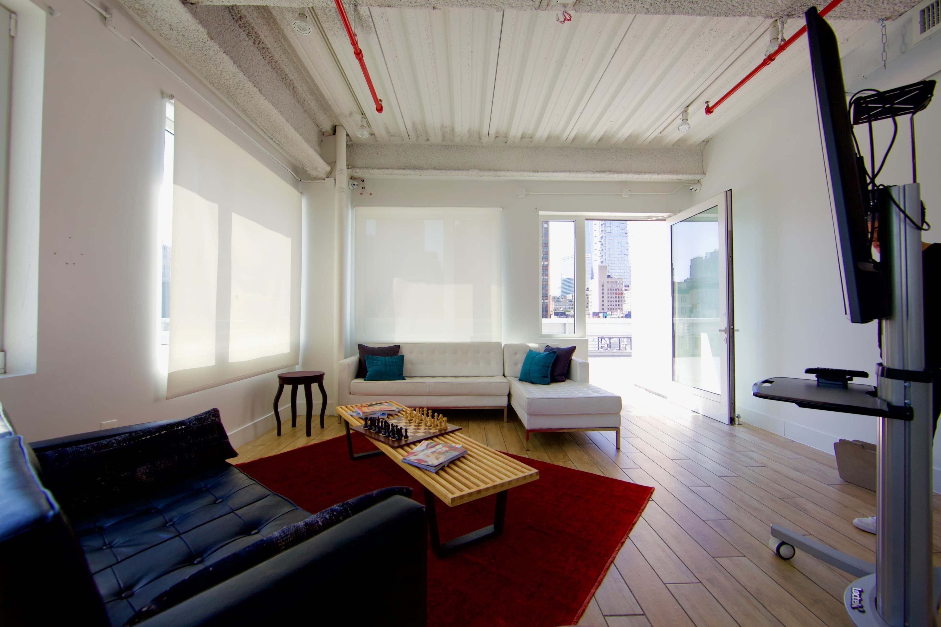A modern, minimalist office lounge area featuring a dark sofa, a white sectional couch, a coffee table with a chess set, and large windows overlooking a cityscape.