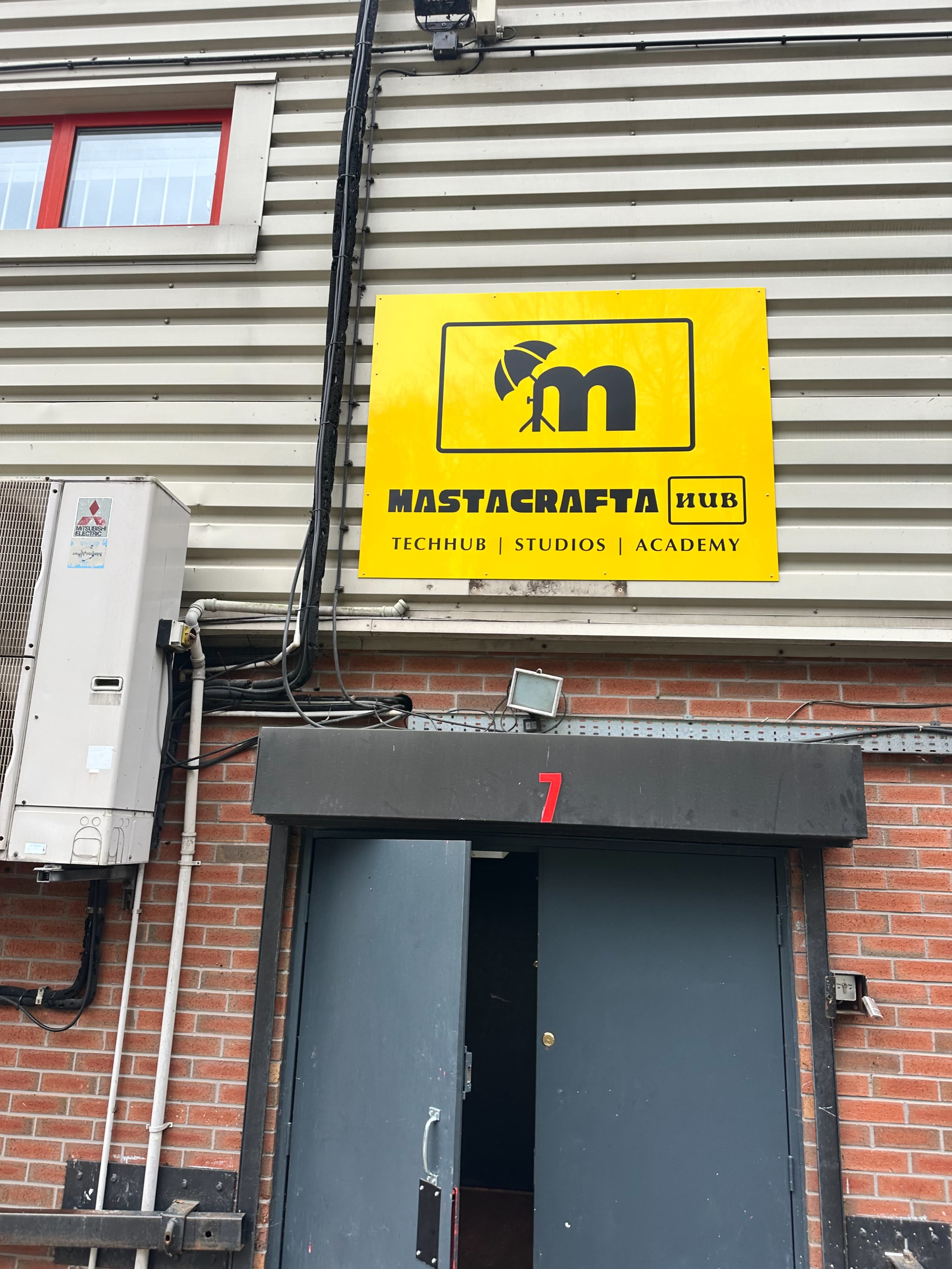 The image shows a building entrance with a large yellow sign displaying the name "Mastacrafta Hub" and its associated labels: "Techhub," "Studios," and "Academy."