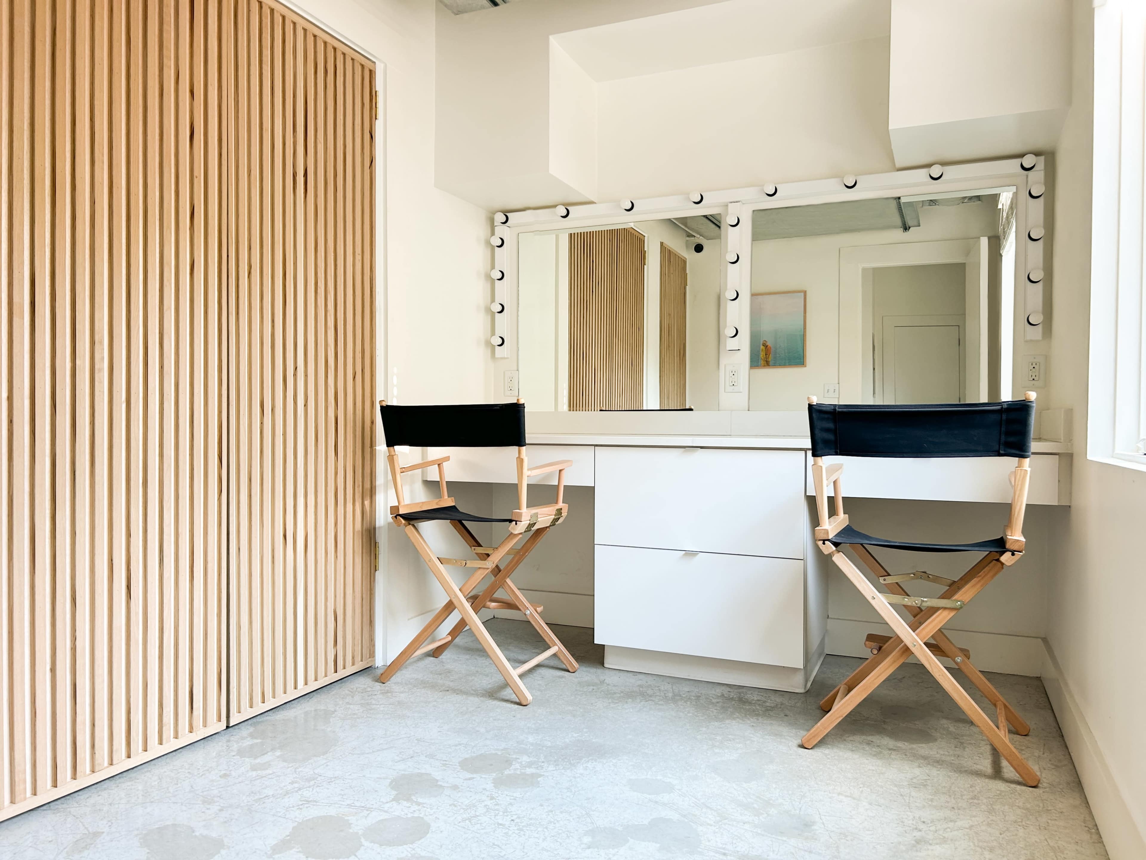 The image shows a well-lit makeup area featuring a large mirror with lights, two director's chairs, and a wooden sliding door.