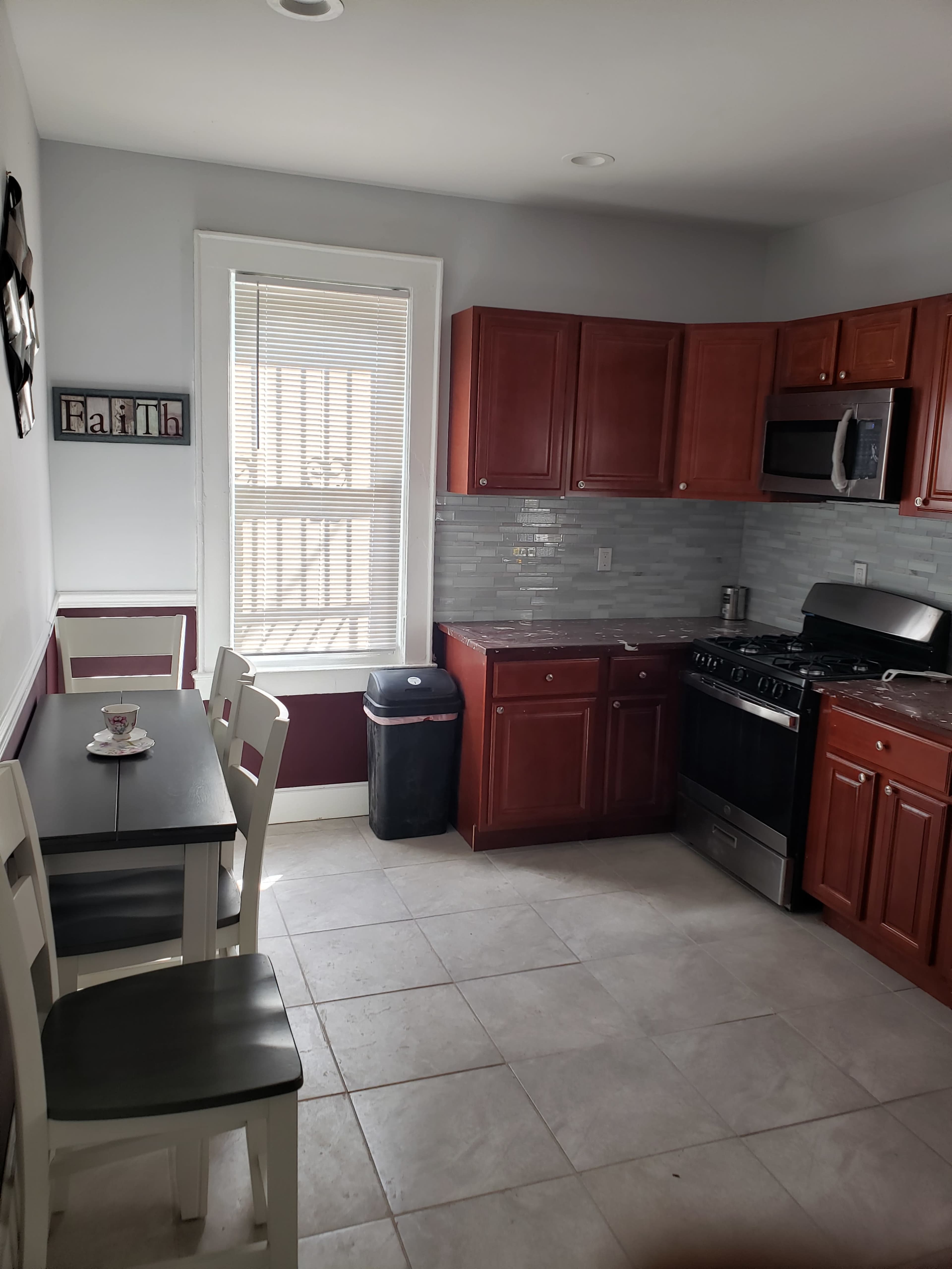 A kitchen features brown wooden cabinets, stainless steel appliances, a small table with chairs, and a window allowing natural light.