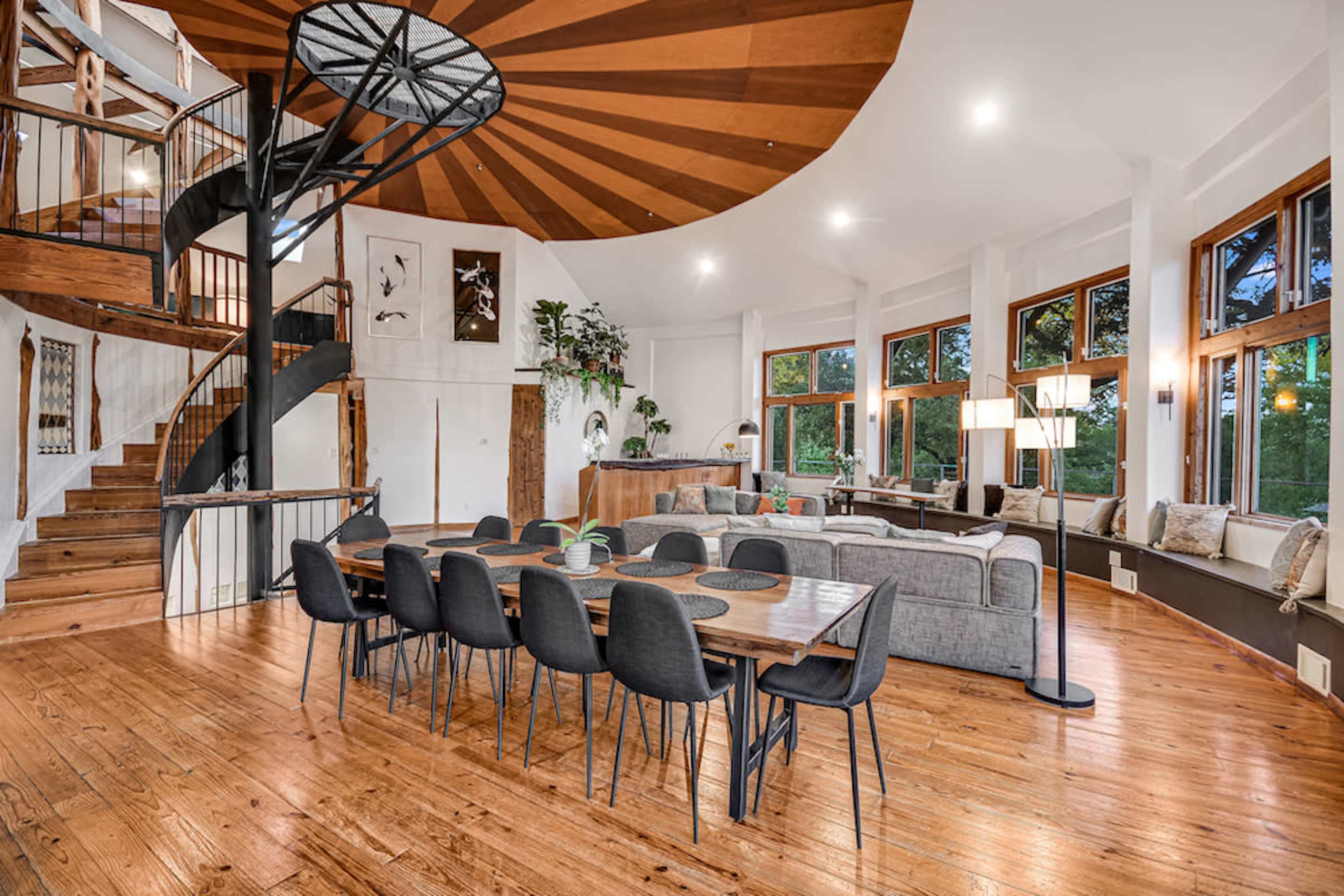 A spacious open-concept living area featuring a large dining table, a spiral staircase, and multiple windows offering natural light.