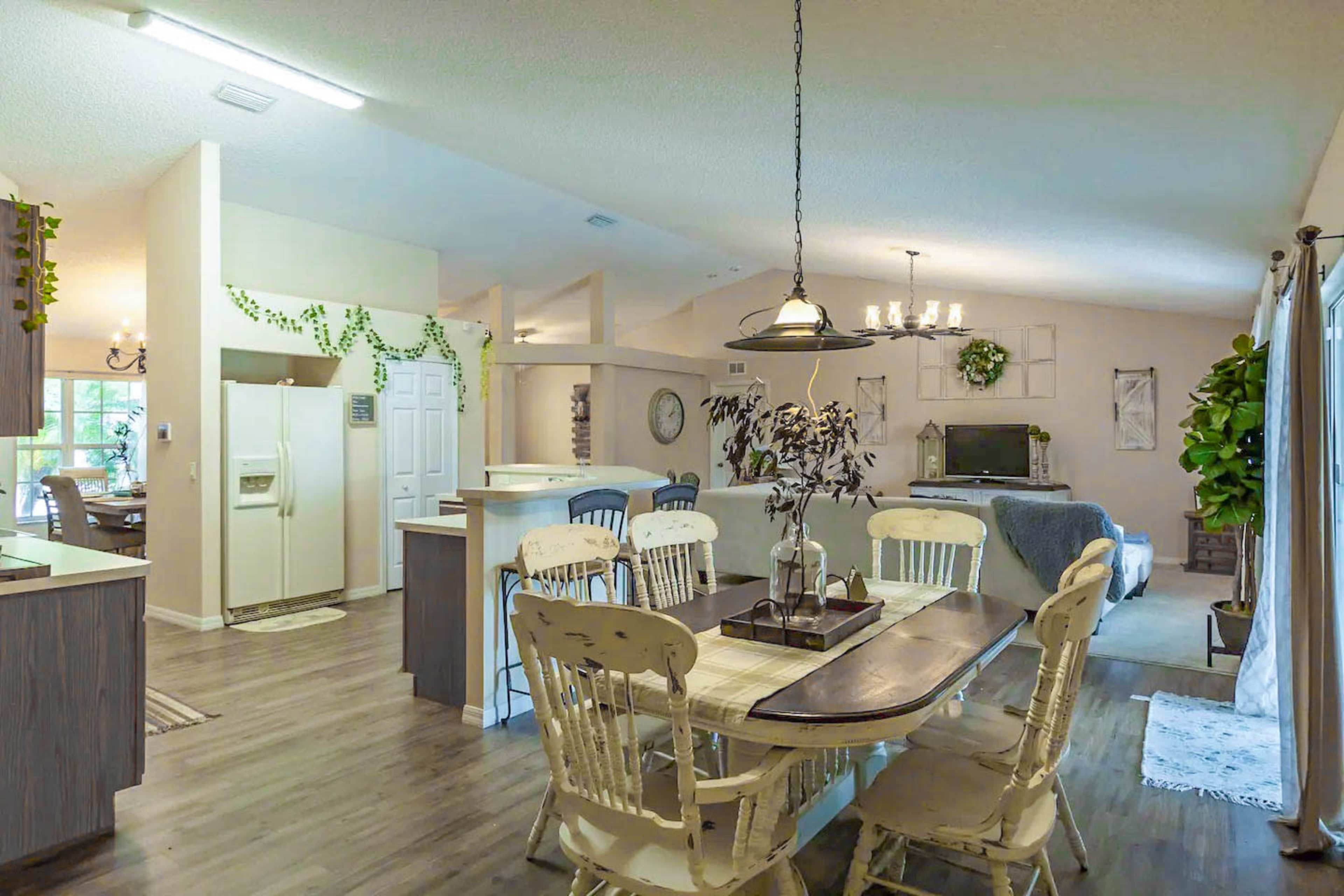 The image shows a spacious interior with a dining area featuring a wooden table and chairs, a kitchen with a refrigerator, and a living room visible in the background.
