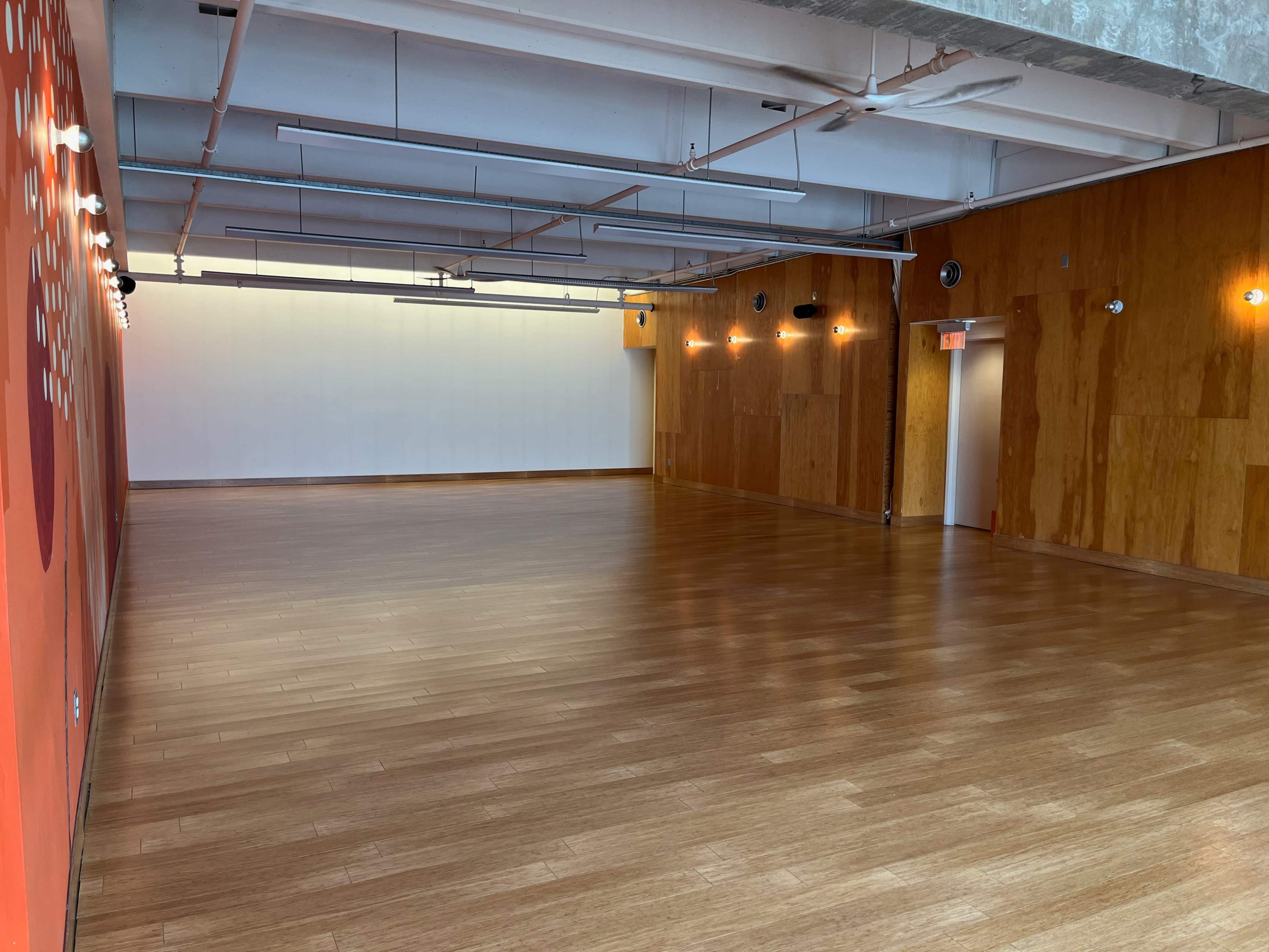 A spacious, empty interior room with wooden flooring and light-colored walls, featuring a few light fixtures along the ceiling.