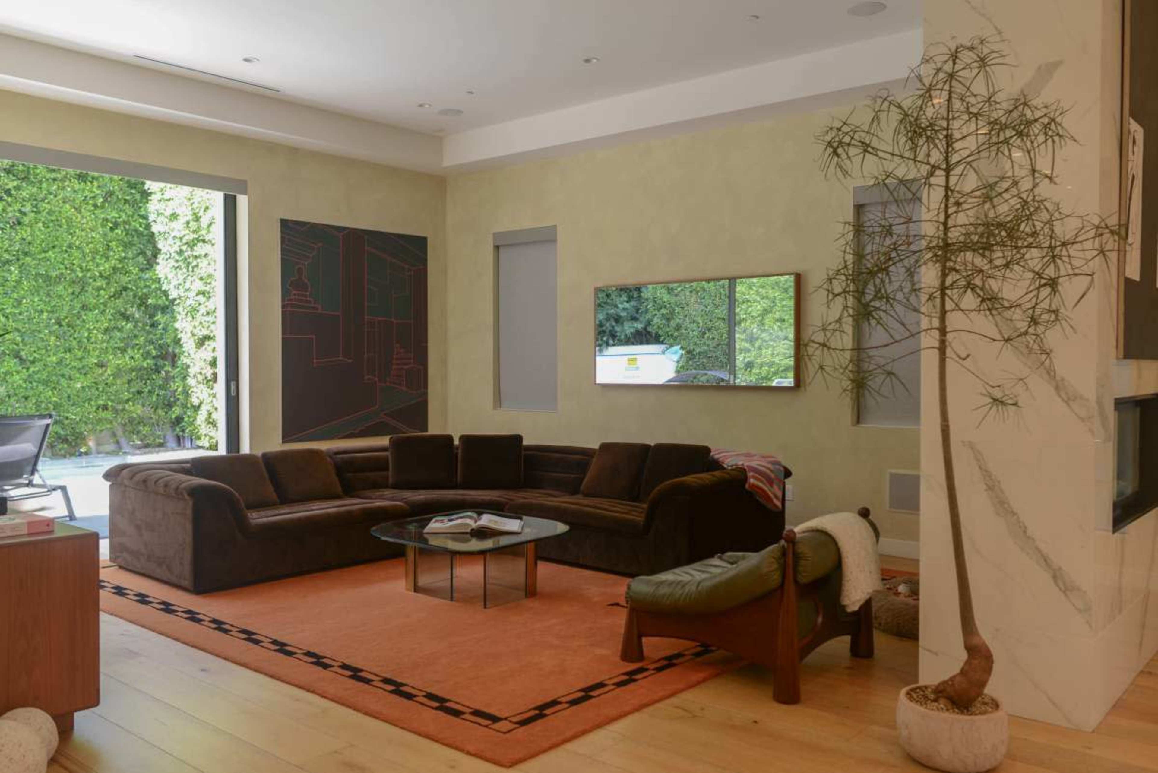 The image shows a modern living room featuring a brown sectional sofa, an orange rug, a glass coffee table, and large windows with greenery visible outside.