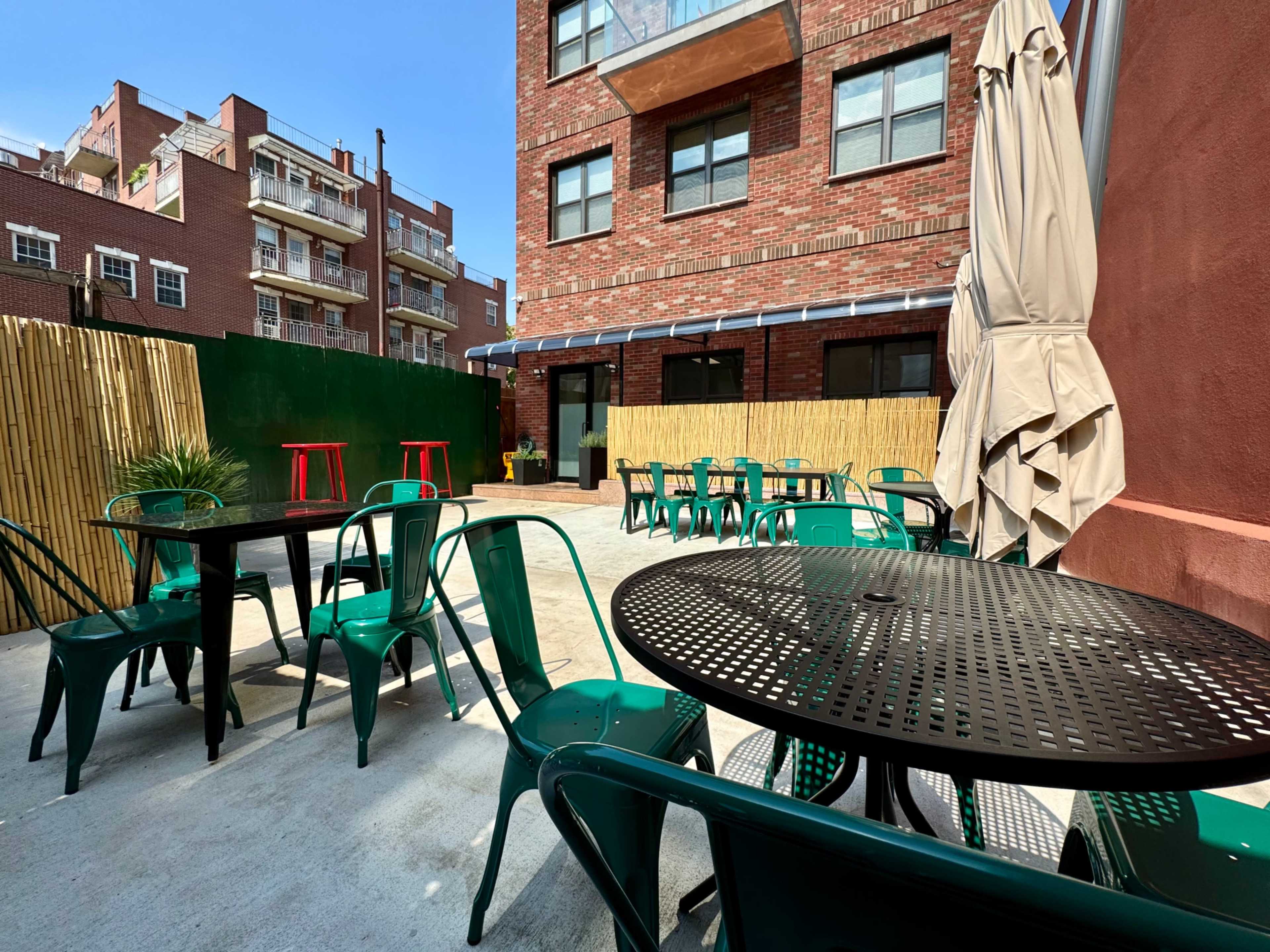 A patio area features green metal chairs and black circular tables, surrounded by brick buildings and a bamboo fence under a clear blue sky.