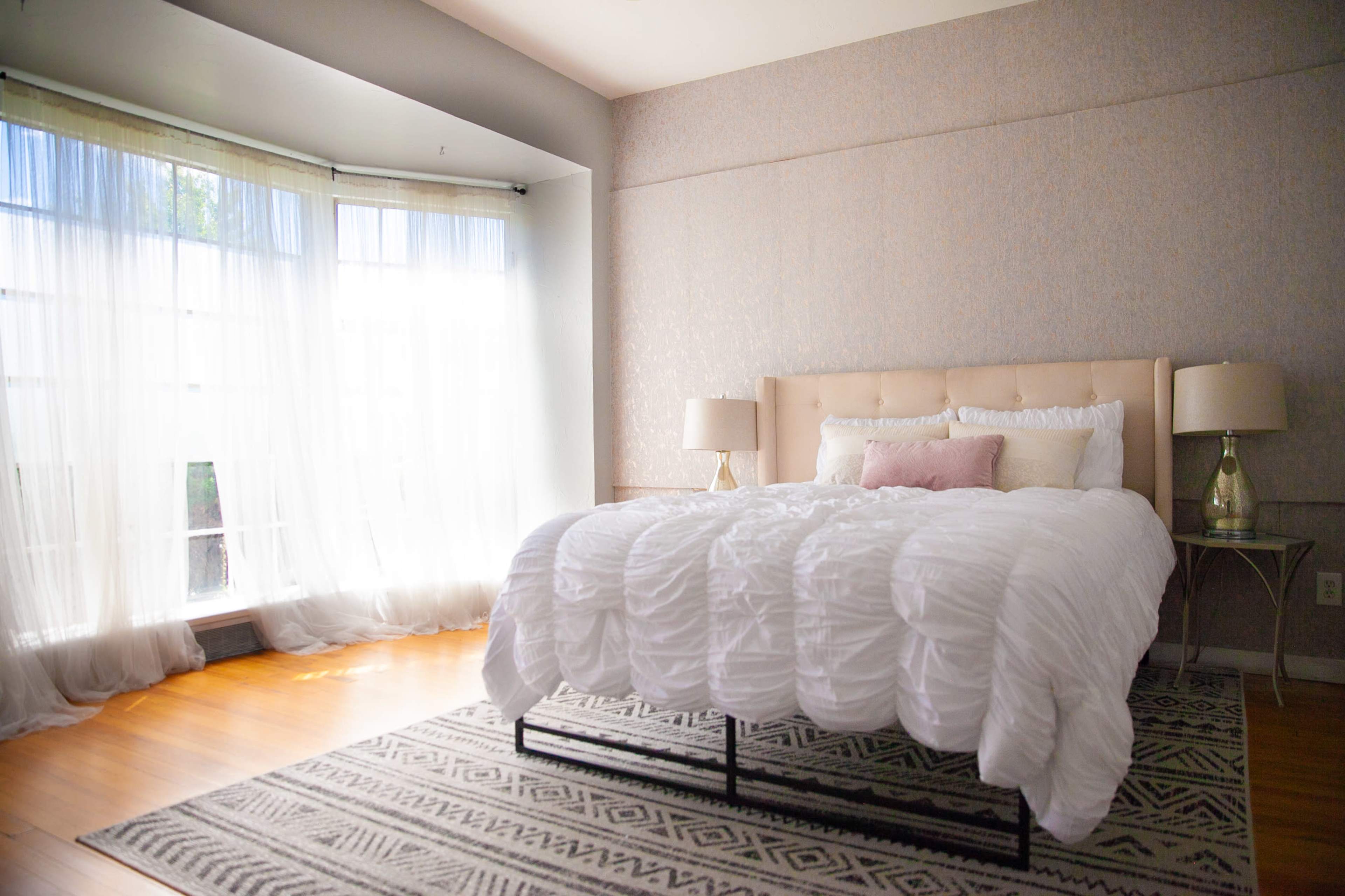 A well-lit bedroom features a bed with a tufted headboard, white bedding, and a patterned rug beneath, with large windows dressed in sheer curtains.