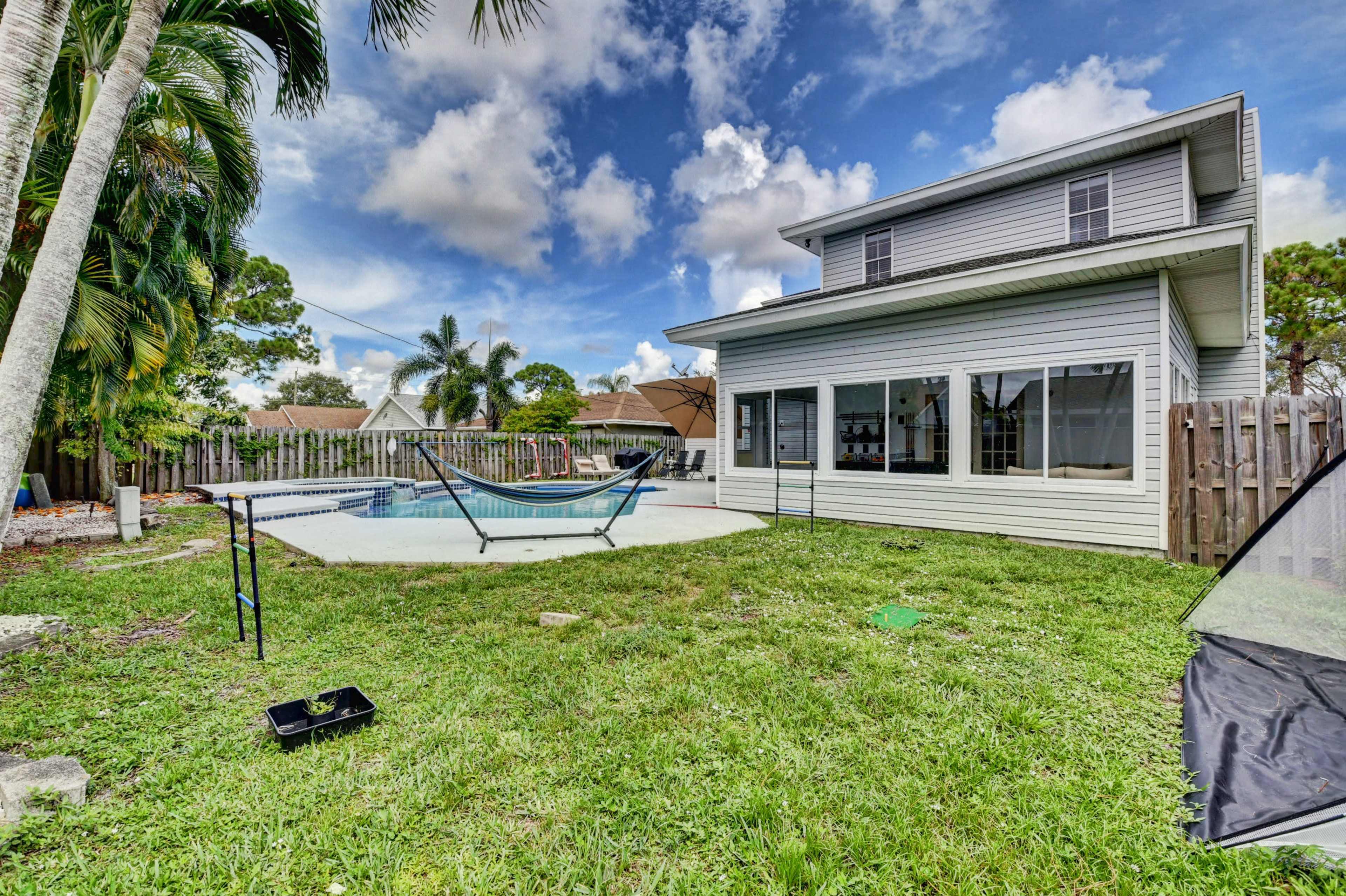 A two-story house with large windows overlooking a backyard featuring a pool, a hammock, and a grassy area surrounded by palm trees.