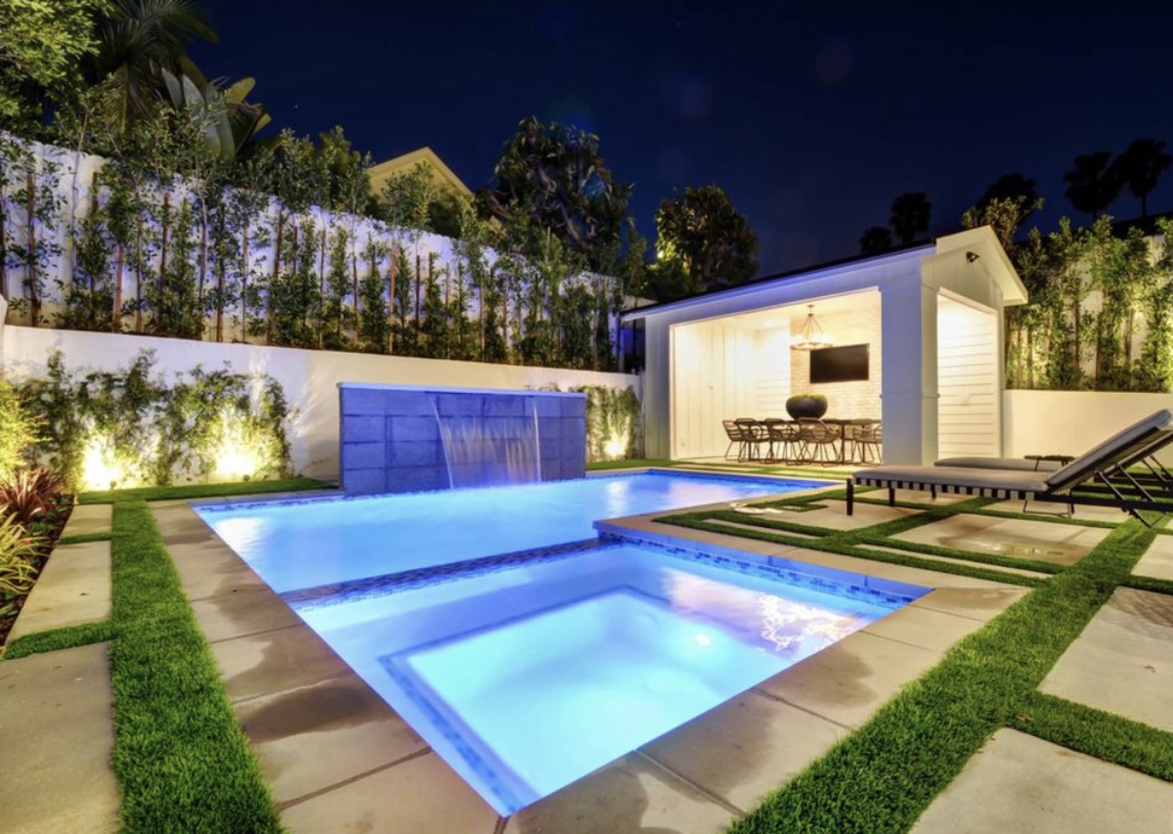 The image shows a modern backyard at night featuring a lit swimming pool with a waterfall, surrounded by green landscaping and a small seating area.