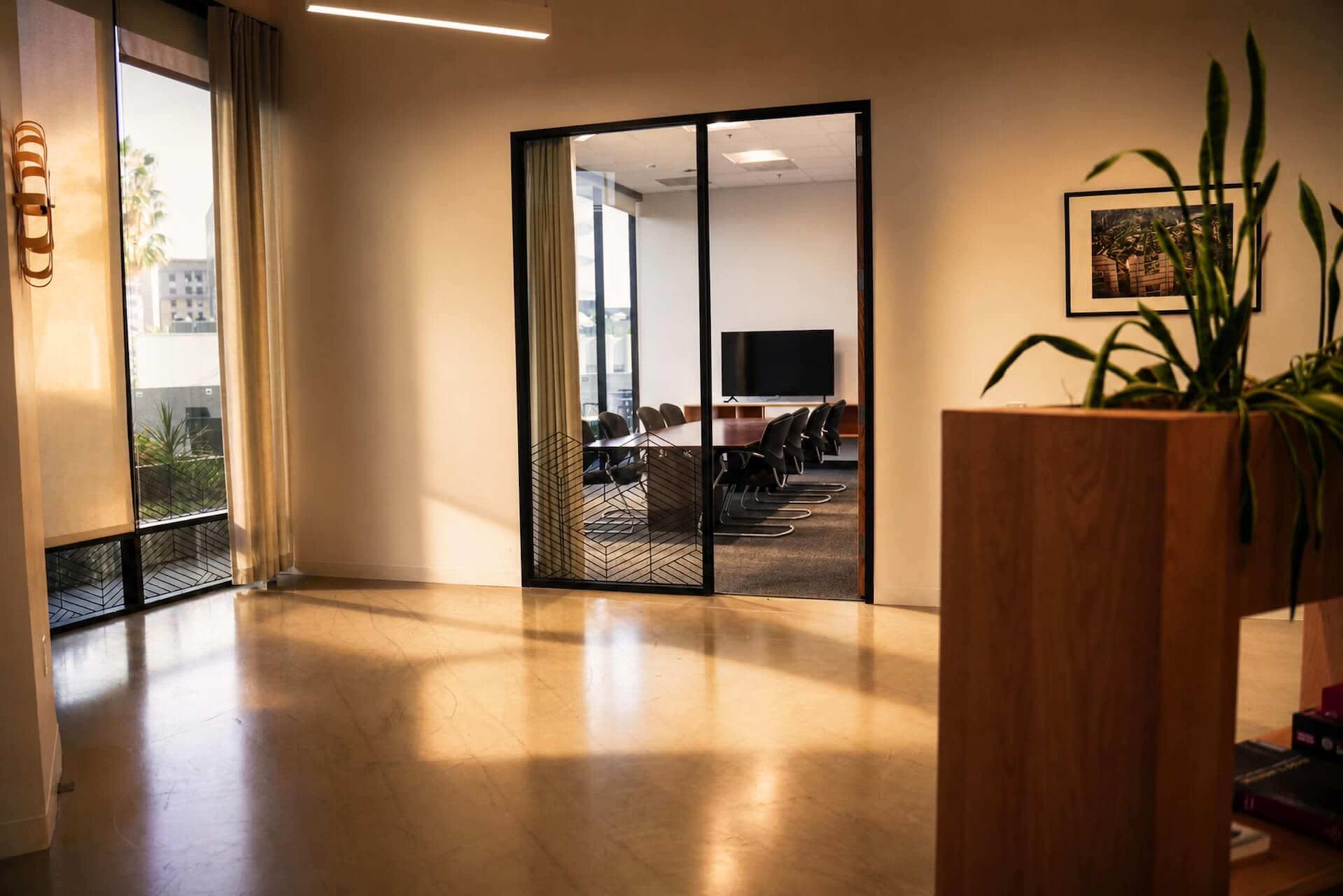 The image shows a modern office space featuring a glass-walled meeting room with a large table and chairs, illuminated by natural light from large windows.