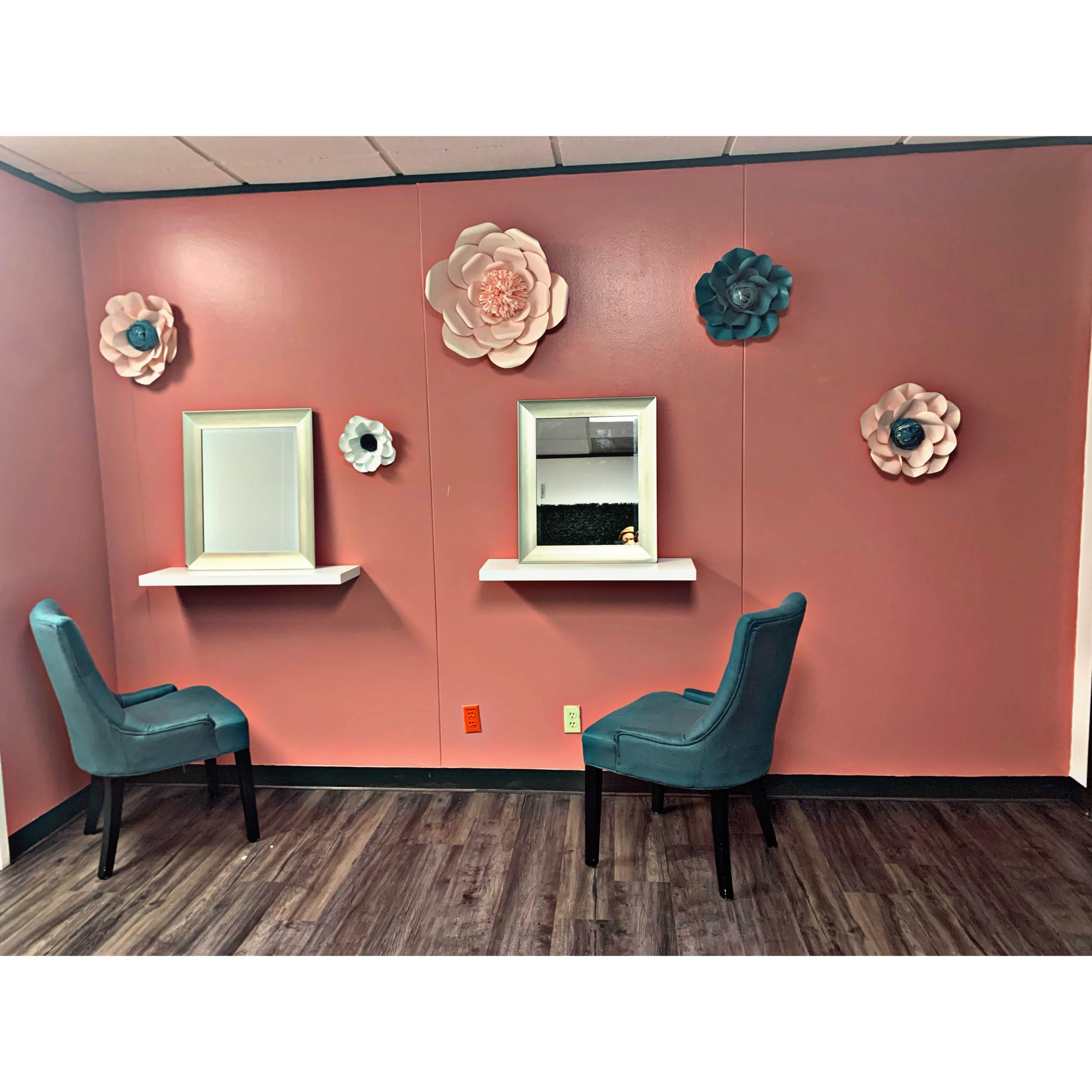 The image shows a room with a pink wall decorated with large floral wall art, two white shelves with mirrors, and two teal chairs facing each other.