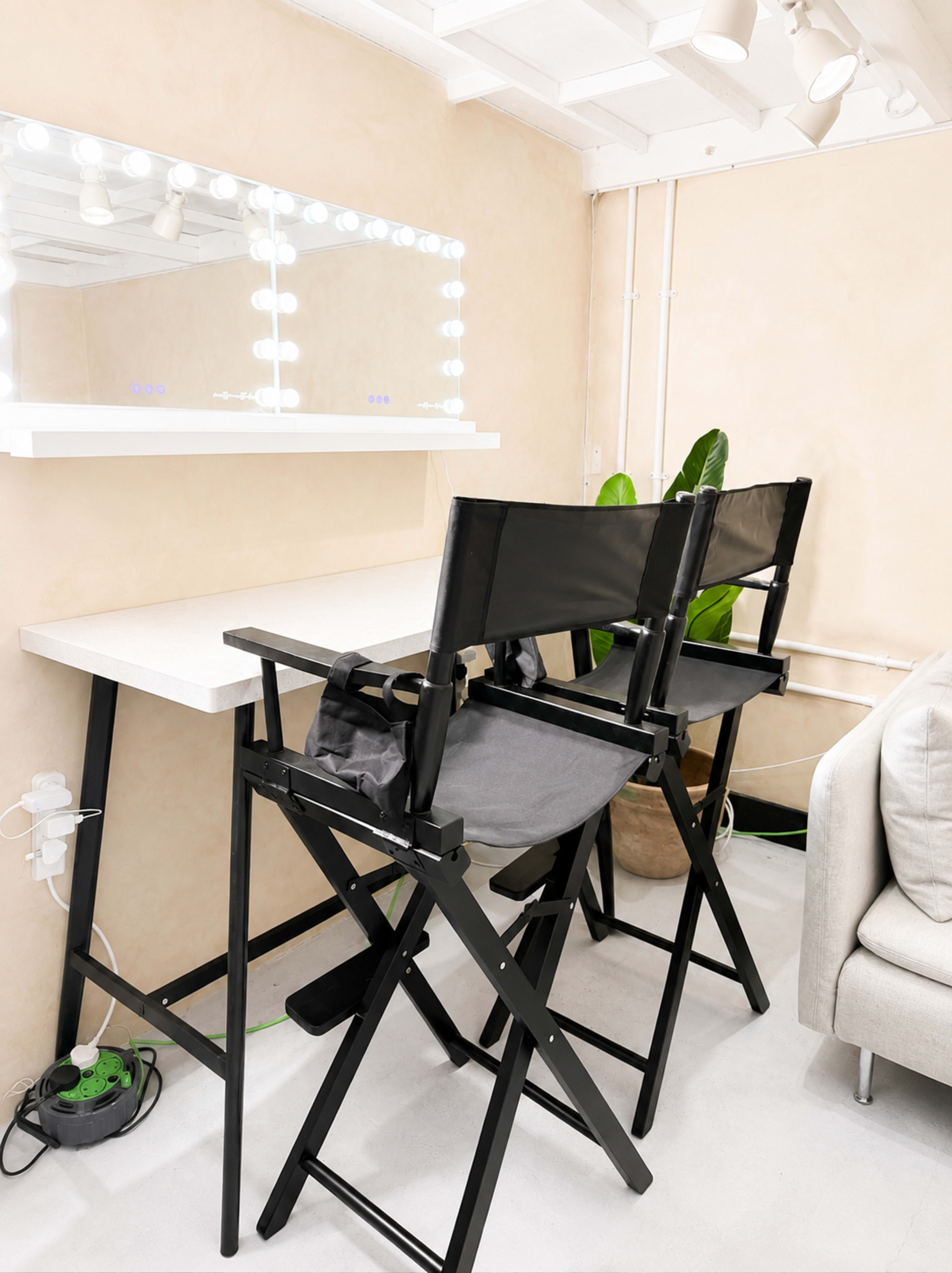A modern makeup station features a large mirror with light bulbs above, complemented by two black director's chairs and a beige wall.