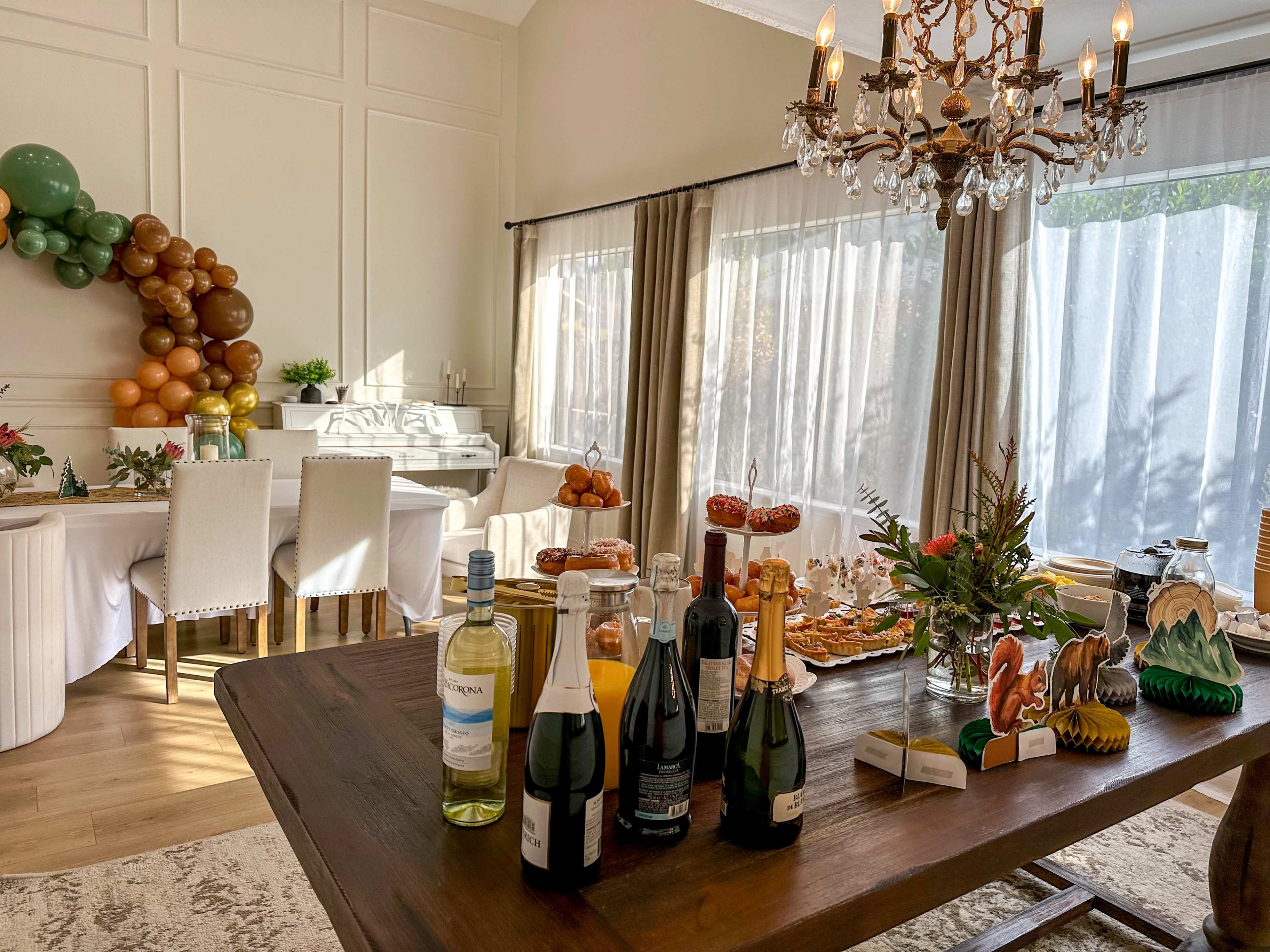 A dining room is set with a variety of food and drinks on a wooden table, featuring decorative balloons and a chandelier overhead.